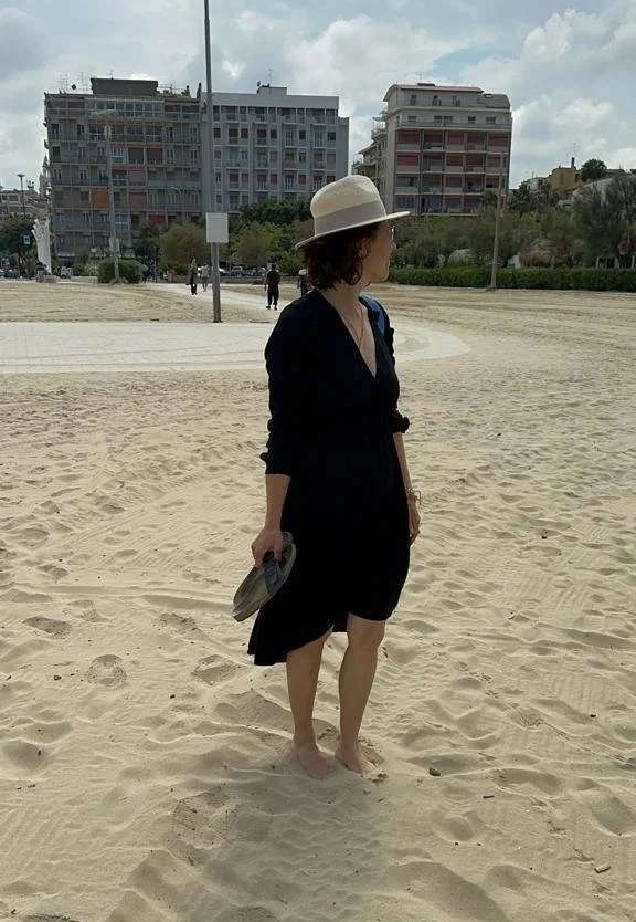 Gaye Gungor in a black dress and wide-brimmed hat standing barefoot on a sandy beach, holding sandals in her right hand, with apartment buildings in the background on a cloudy day.