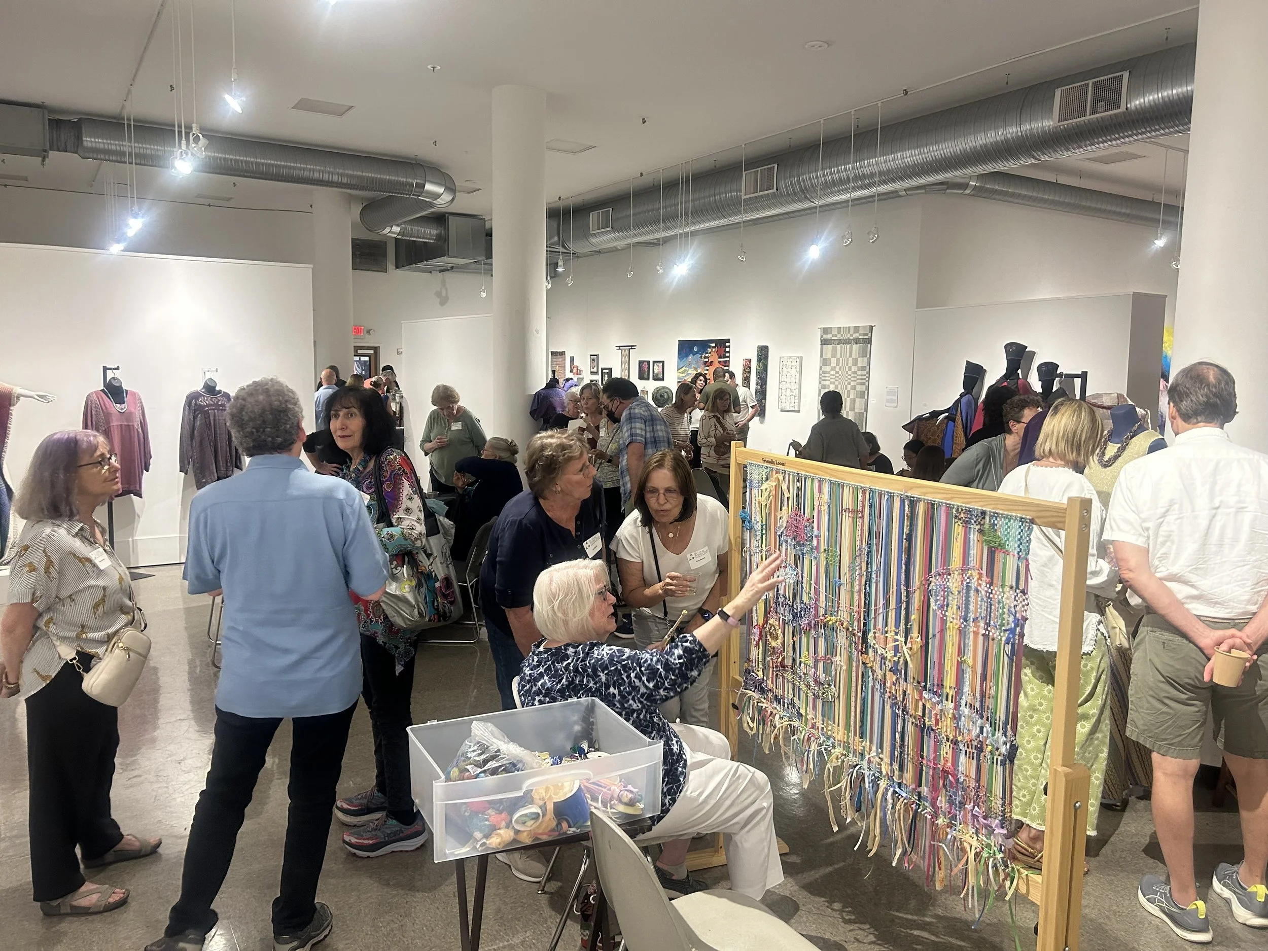 People attending an art or craft exhibition, browsing colorful handmade bracelets displayed on a wooden stand, with various art pieces on the walls and mannequins dressed in clothing in the background.