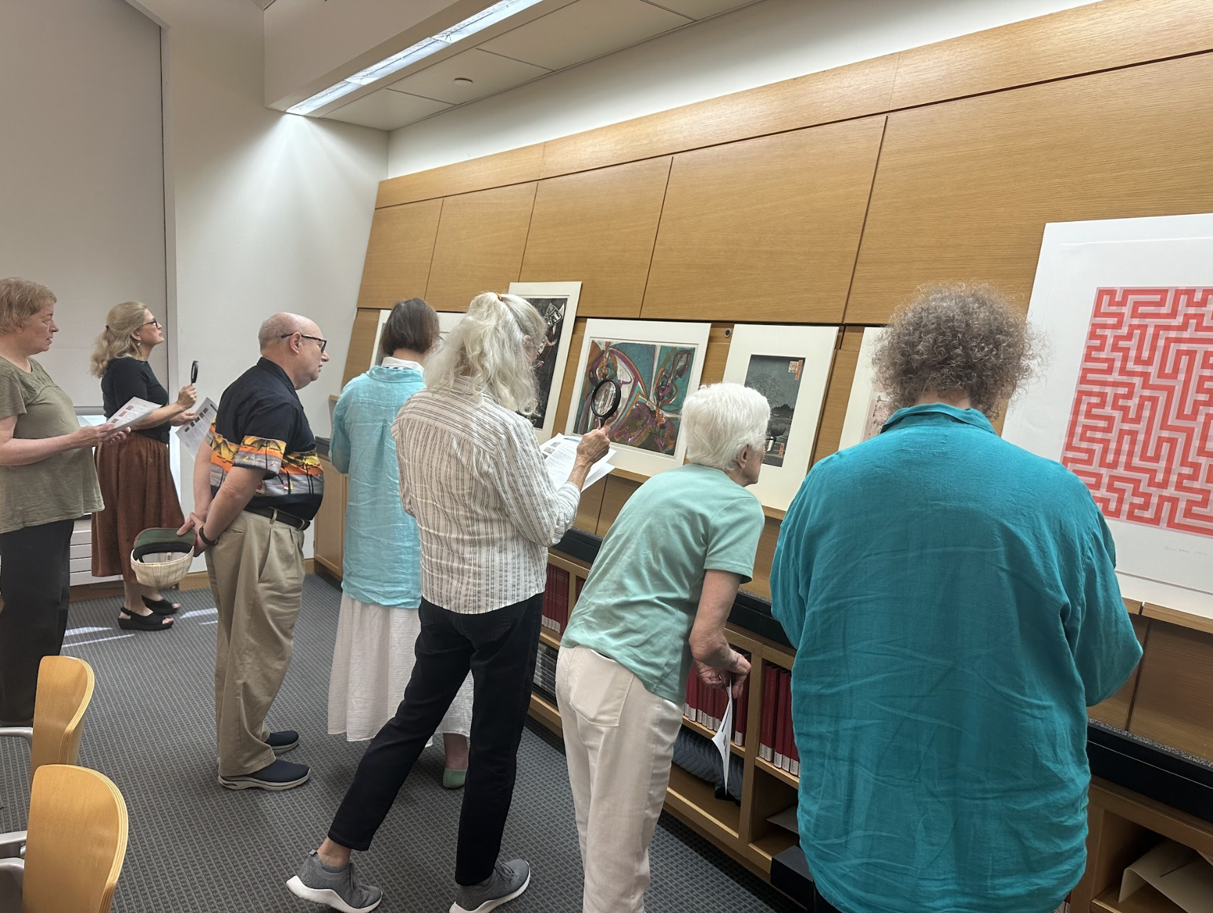 A group of elderly people observing art pieces on display at The Rare Print Room at the St. Louis Art Museum. Some are holding pamphlets, one is holding a magnifying glass, and they are looking closely at the framed artworks on the wall.