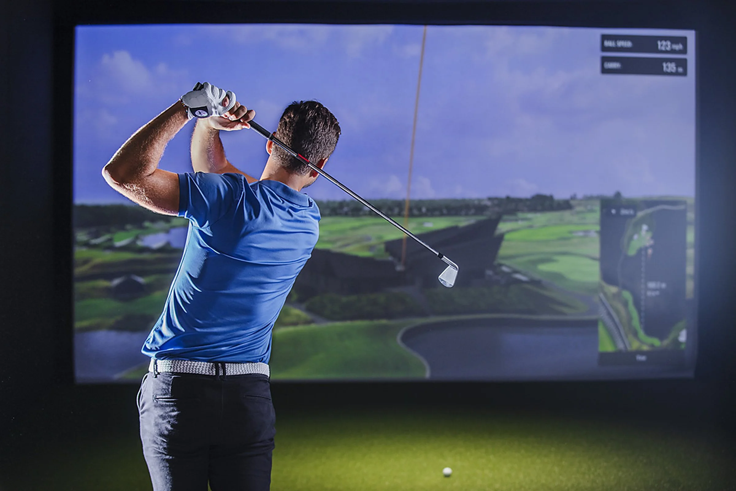 A man in a blue golf shirt and black pants is swinging a golf club on an indoor golf simulator. The background shows a digital golf course with green fairways and water hazards.