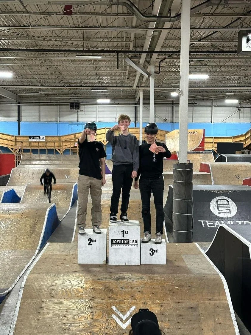 Three young individuals standing on a winners' podium at an indoor skate park, holding medals and certificates after a skating competition. The middle person is in 1st place, the left in 2nd, and the right in 3rd, with a dirt ramp and skateboarding area in the background.