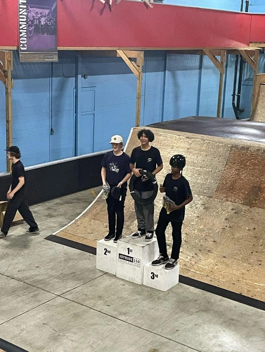 Three children standing on a winners' podium at an indoor skate park, with the first-place child holding a helmet and a skate, second-place child holding a bottle, and third-place child holding a paper; skateboard ramps and a blue wall are visible in the background.