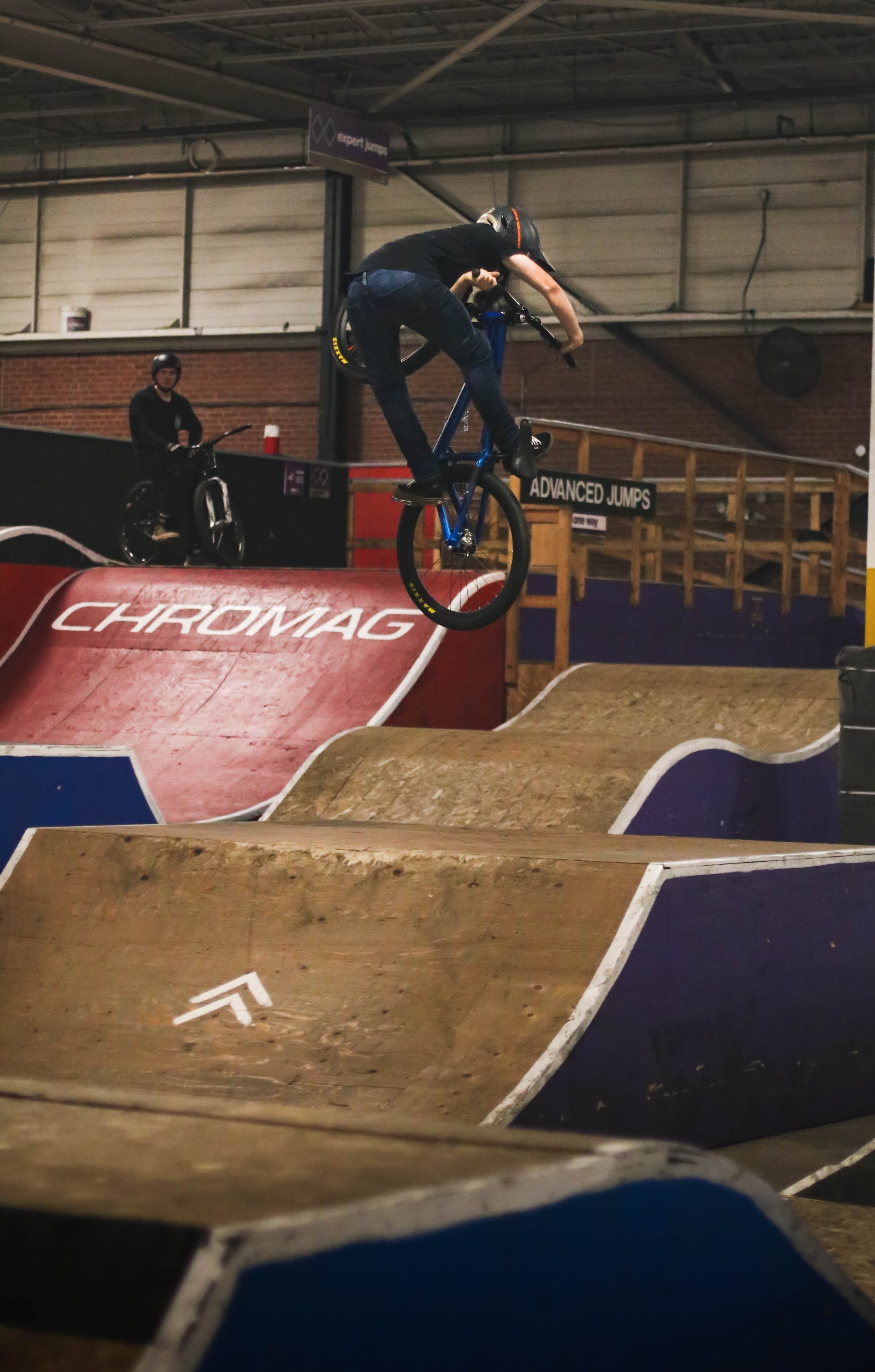 A person performing a jump on a BMX bike at an indoor skate park, with another person watching nearby.