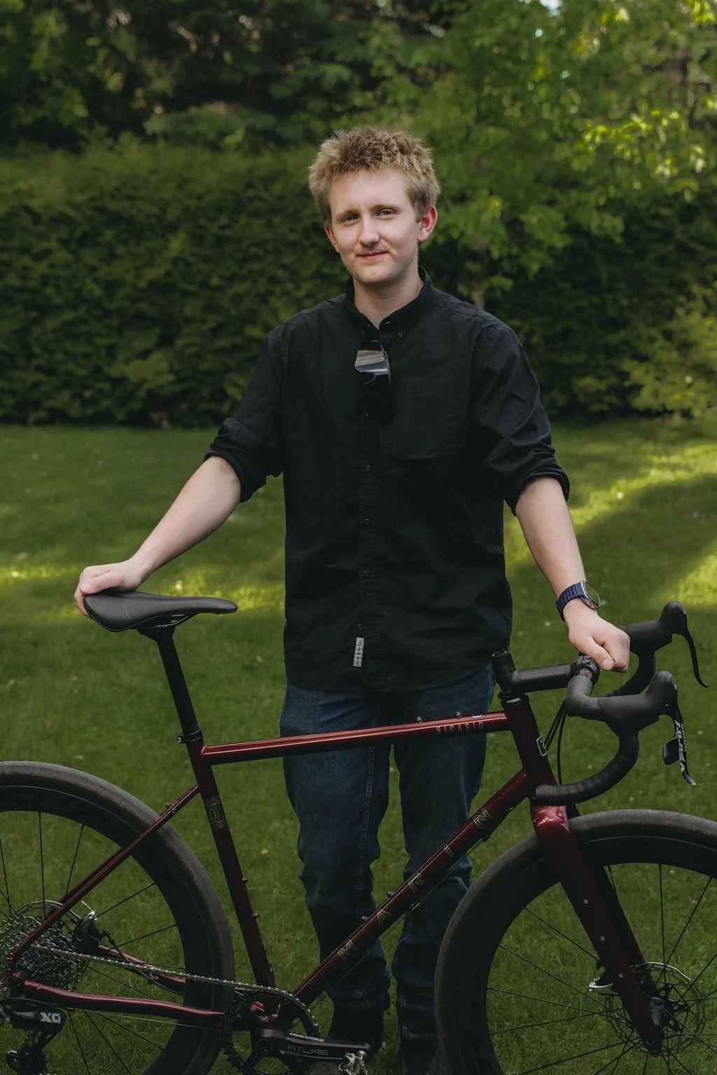 A young man with light-colored, messy hair, wearing a black button-up shirt with sleeves rolled up, posing outdoors with a red bicycle on a grassy area with trees in the background.