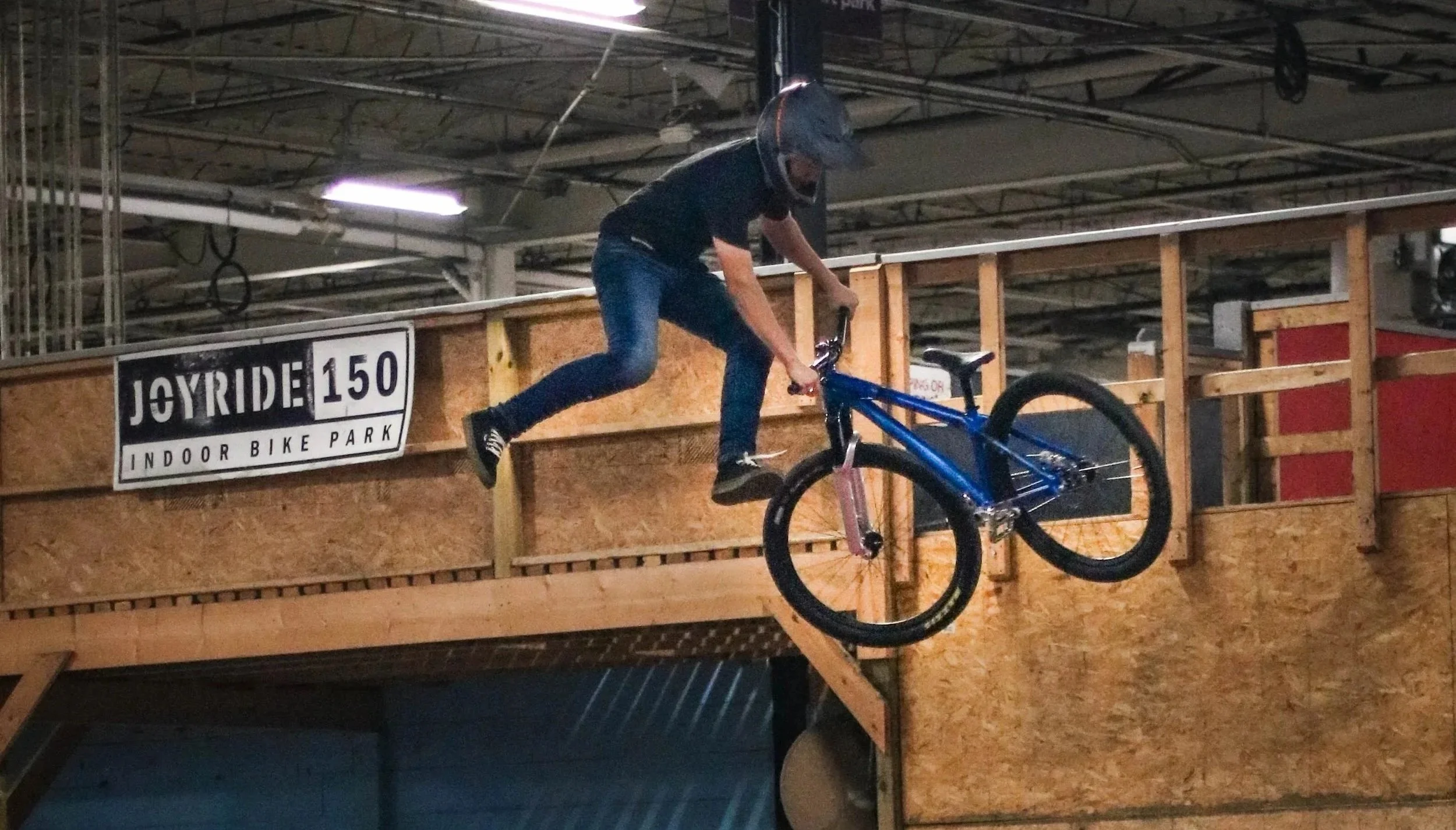 Person wearing a helmet riding a BMX bike at an indoor skate park, jumping off a ramp near a sign that says 'Joyride 150 Indoor Bike Park'.