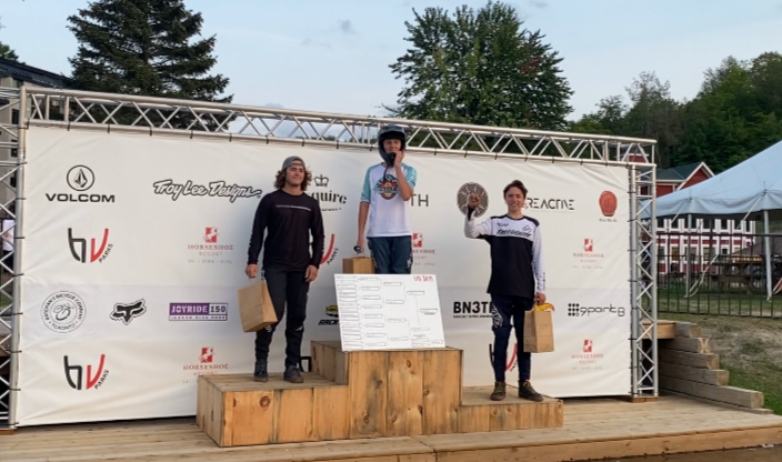 Three young men on a wooden podium with a backdrop of sponsor logos, celebrating a competition victory outdoors during daytime.