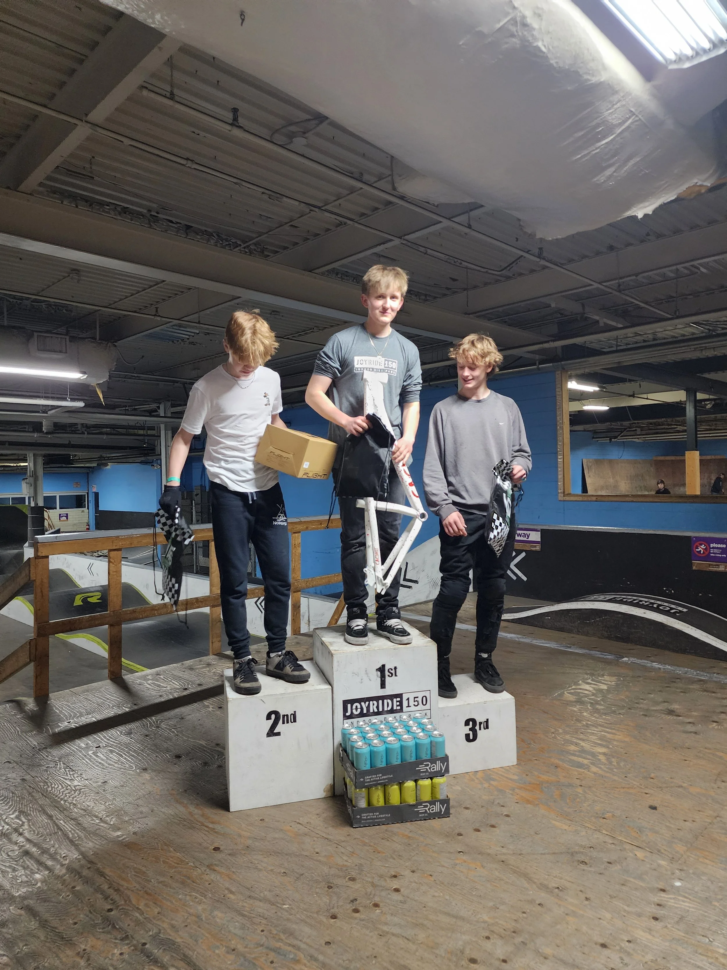Three boys standing on a podium at a skatepark, with the boy in the middle on the first place spot, holding a black cloth. The boy on the left stands on the second place podium and the boy on the right on the third. They are smiling and holding checkered flags. There are stacks of canned drinks at the foot of the podiums, and skate ramps and a blue wall in the background.