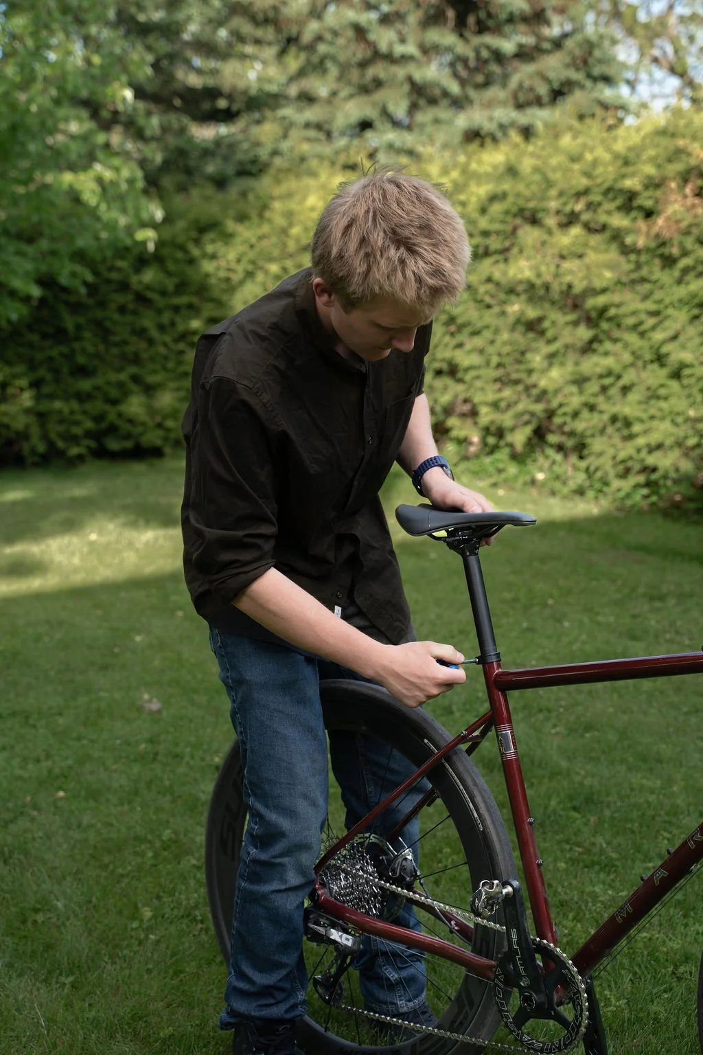 A young man with blond hair wearing a black shirt and jeans is fixing a red mountain bike outdoors on a grassy area with green foliage in the background.
