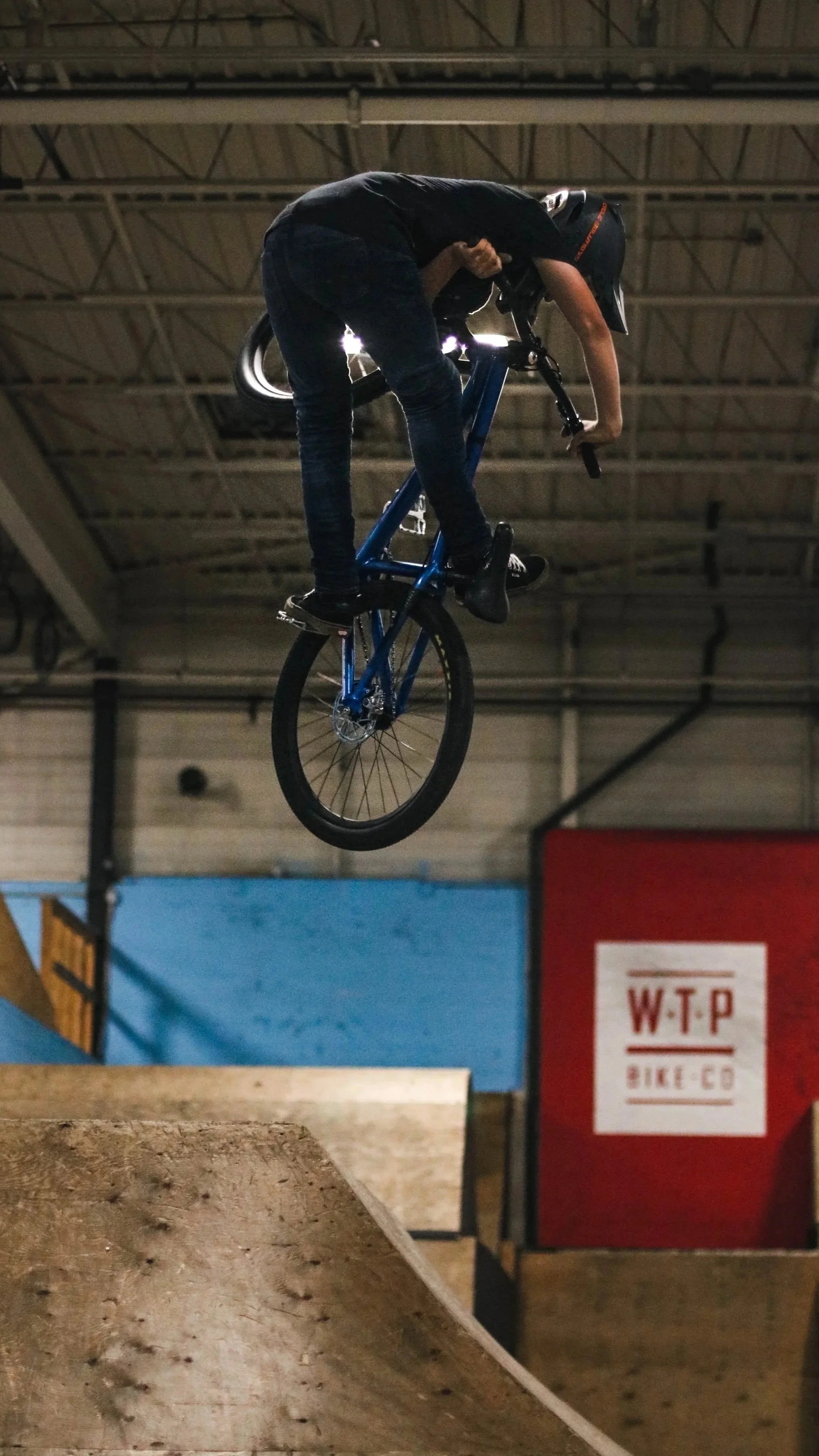A person wearing a black helmet performing a trick on a blue BMX bike at an indoor skatepark.