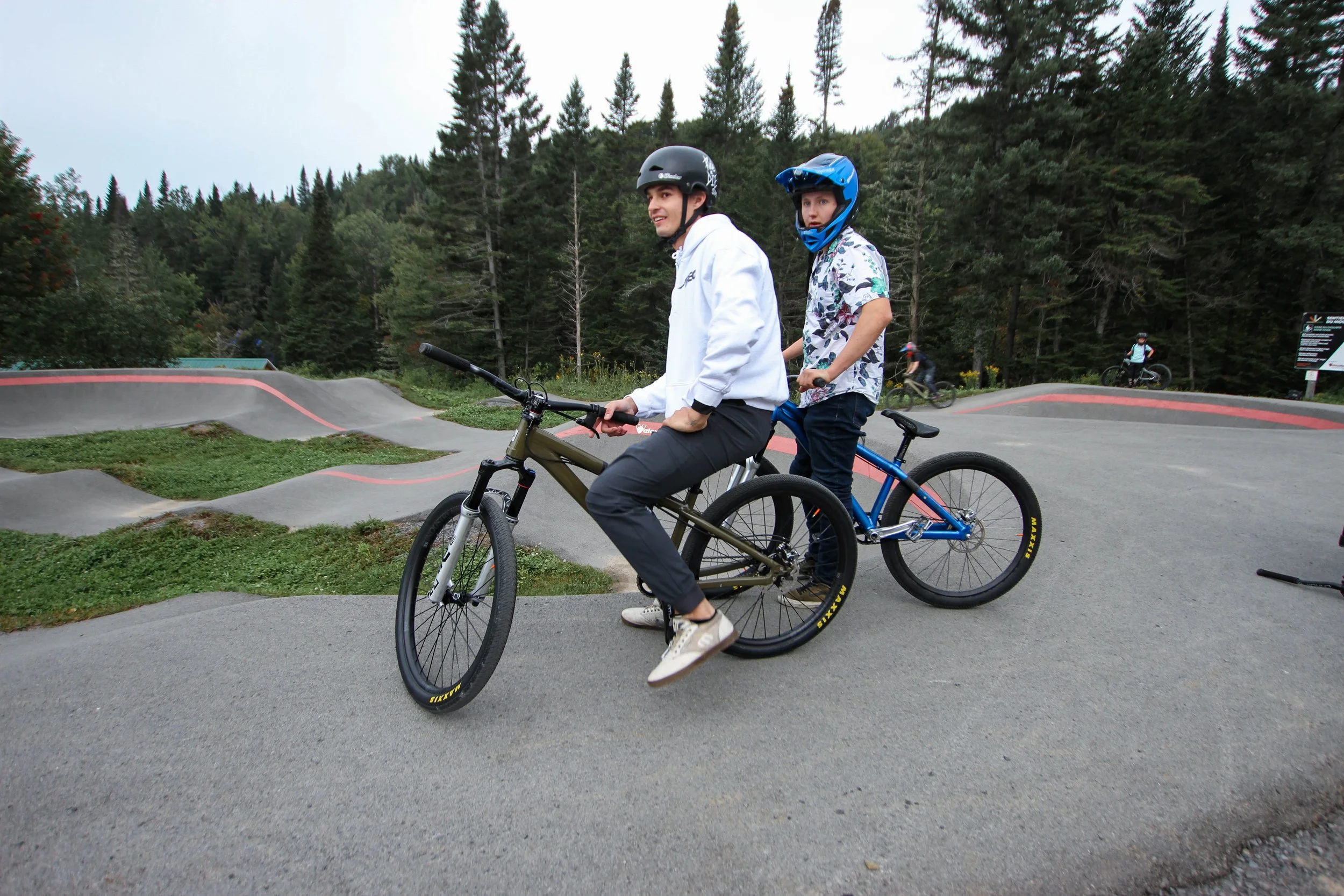 Two young men with helmets riding mountain bikes on a dirt pump track surrounded by trees.