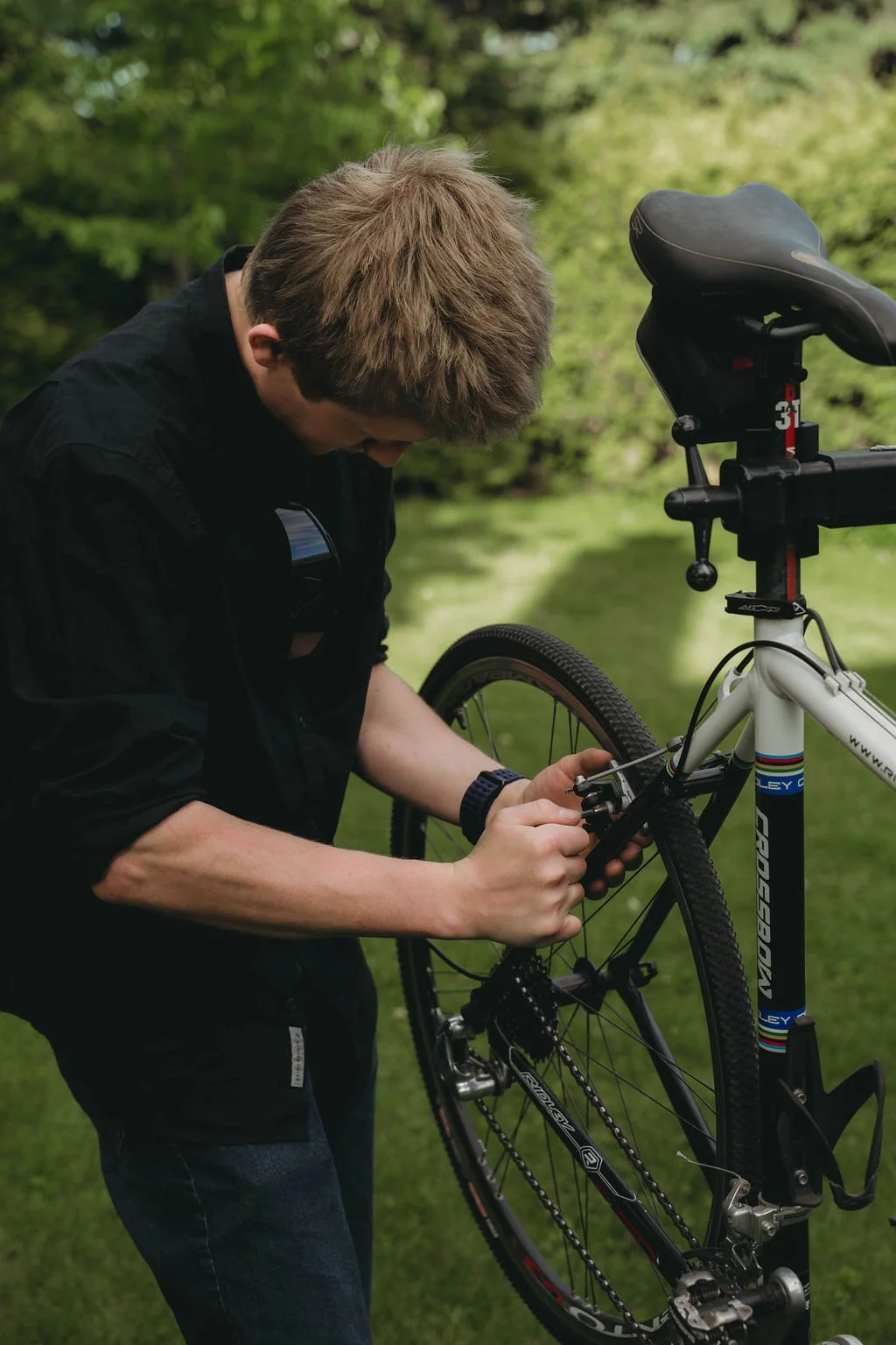 Person repairing a bicycle outdoors near a body of water with green trees in the background