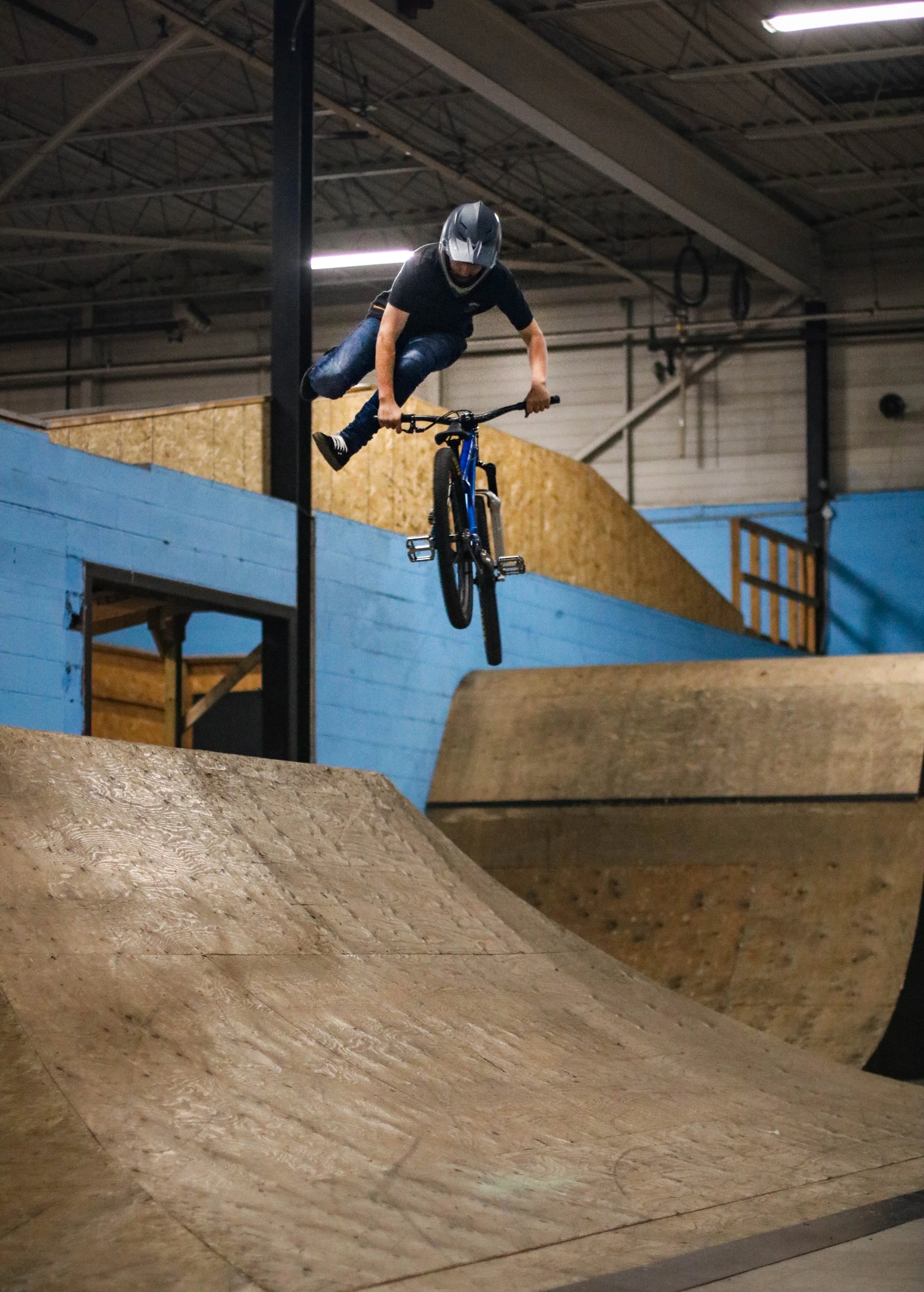 Person wearing a helmet riding a bicycle in an indoor skatepark with wooden ramps and blue walls.