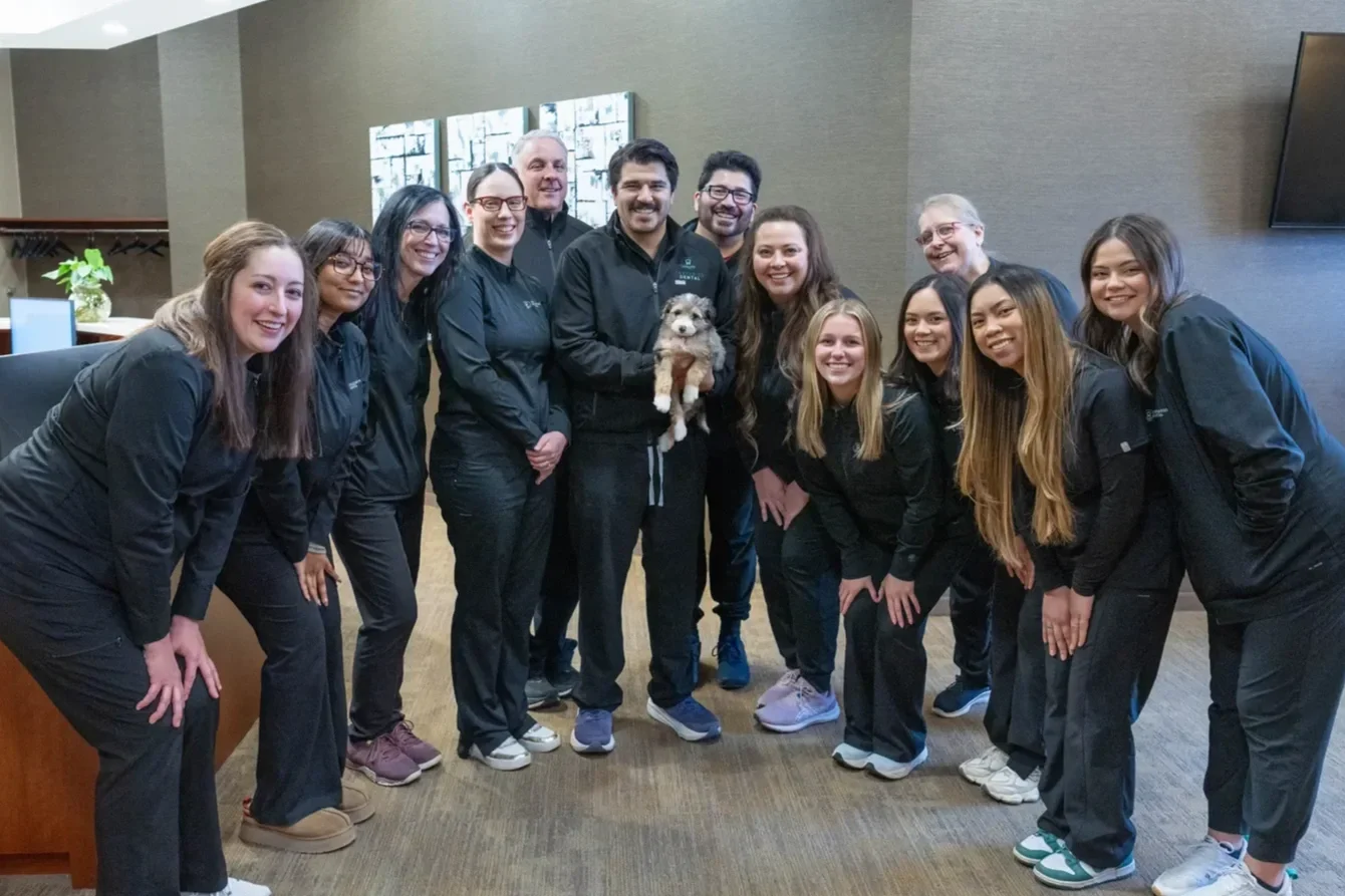 Group of eleven smiling people in black uniforms, including a man holding a puppy, gathered in a room with gray walls and wood flooring.