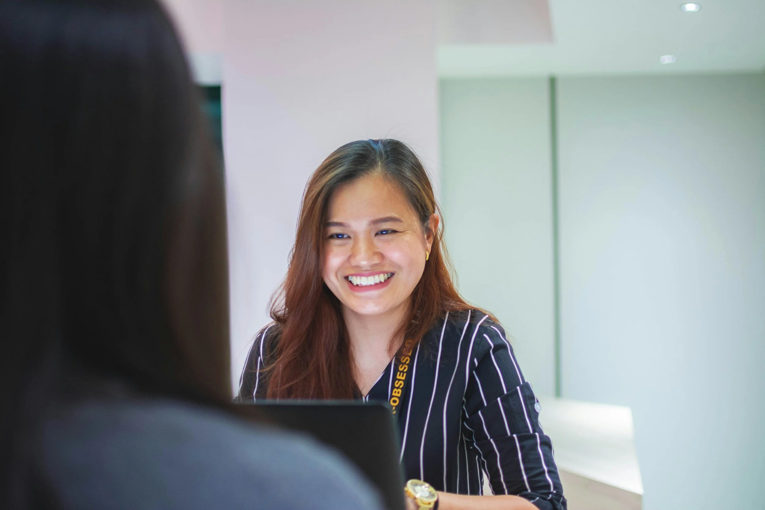 A woman with long brown hair smiling while talking to someone, wearing a black and white striped shirt and a lanyard, in an indoor setting with light-colored walls.