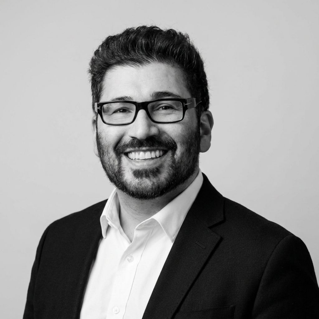Black and white professional headshot of a man with dark hair, beard, glasses, wearing a suit and white shirt, smiling against a plain background.