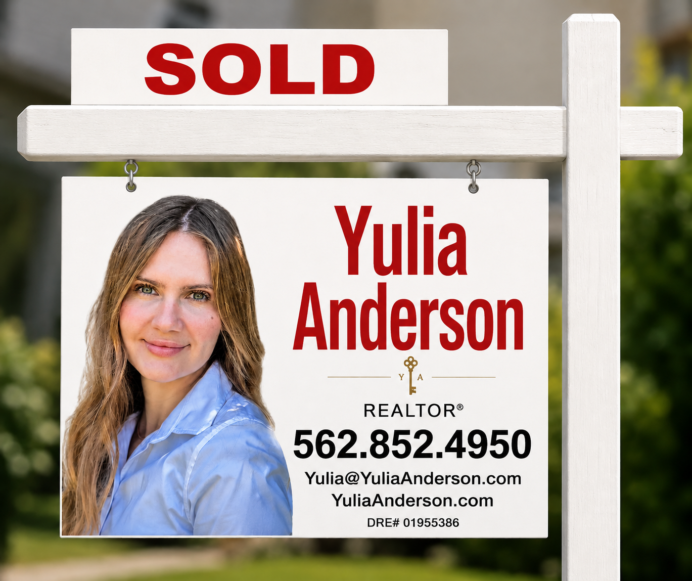 A real estate sign featuring a-photo of a woman with long, light brown hair, wearing a light blue shirt. The sign indicates property is sold, displaying the name Yulia Anderson, with a contact phone number, email, and website.