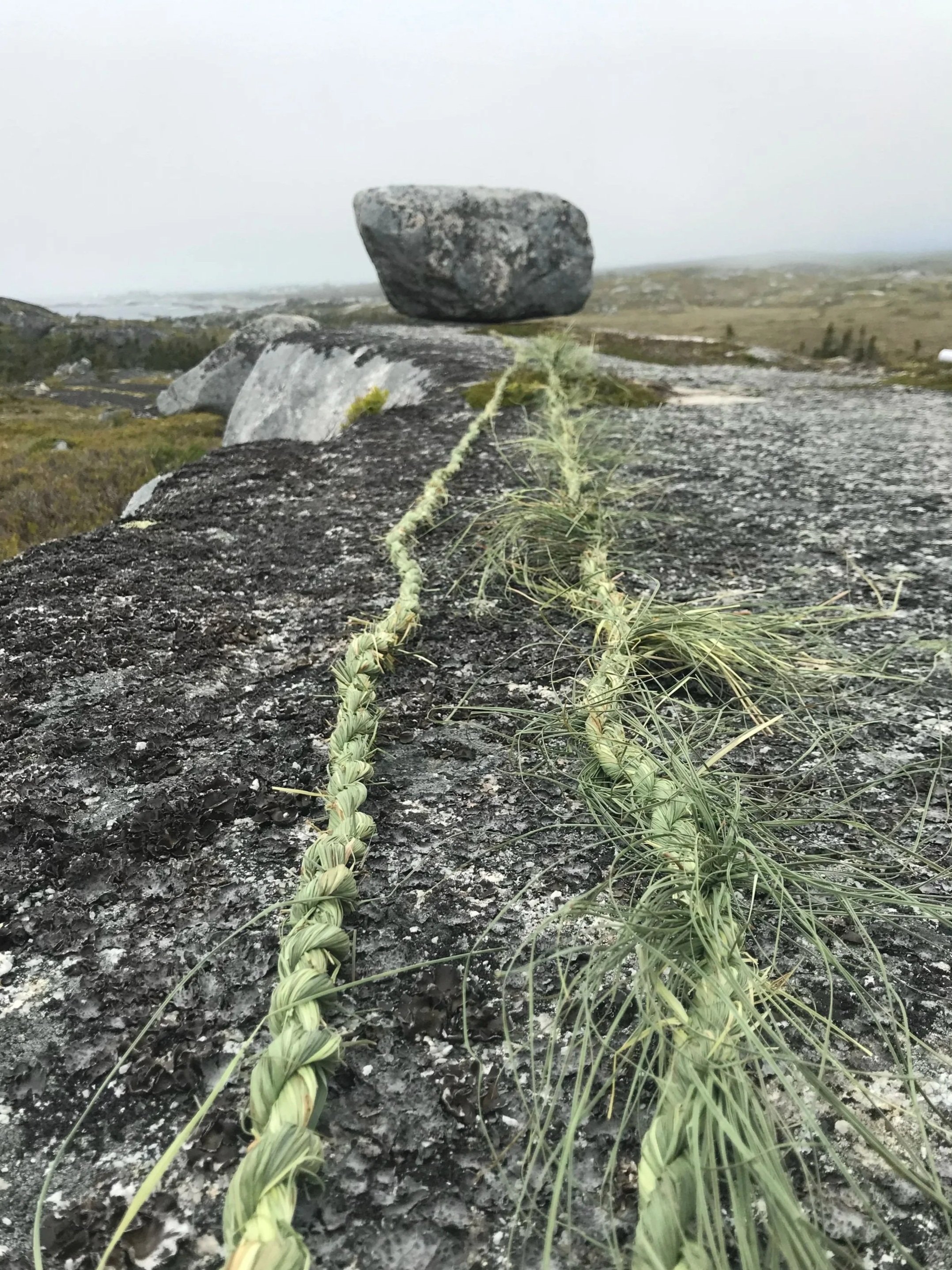 A large boulder balanced on top of smaller rocks in an open, grassy landscape under a cloudy sky.