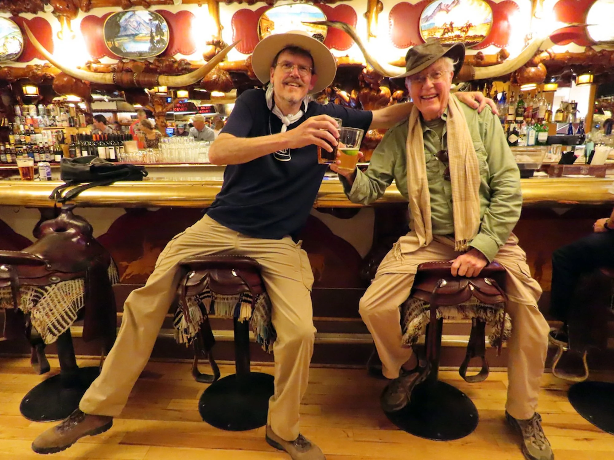 David and father Bob Lauterborn sit in the saddle stools at the Million Dollar Cowboy Bar in Jackson, Wyoming, raising glasses in a toast, with a background decorated with mounted animal horns and paintings, and a lively bar scene behind them.