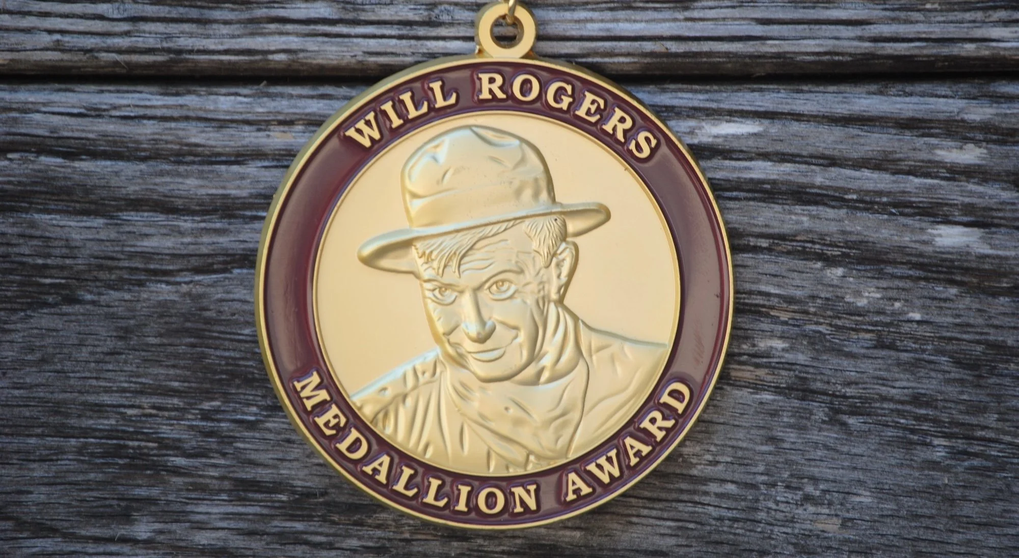 A circular medal featuring a young boy wearing a wide-brimmed hat, with the words 'WILL ROGERS MEDALLION AWARD' inscribed around the circumference, placed on a weathered wooden surface.