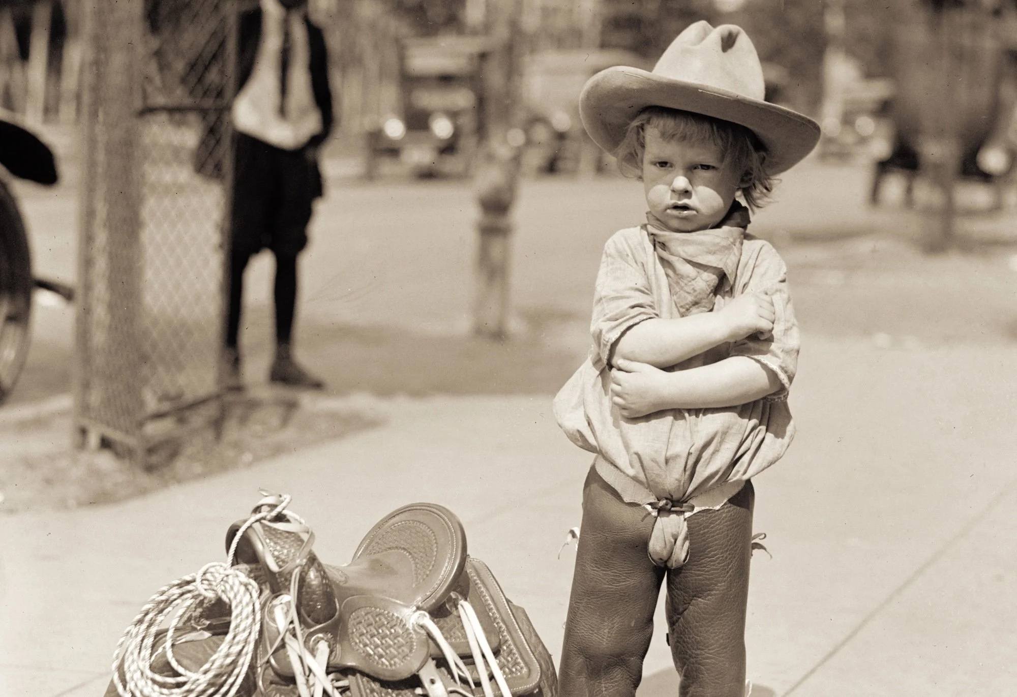 A young boy with curly hair wearing a cowboy hat and bandana, standing with arms crossed next to a basket filled with shoes and ropes, outdoors with blurred figures in the background.