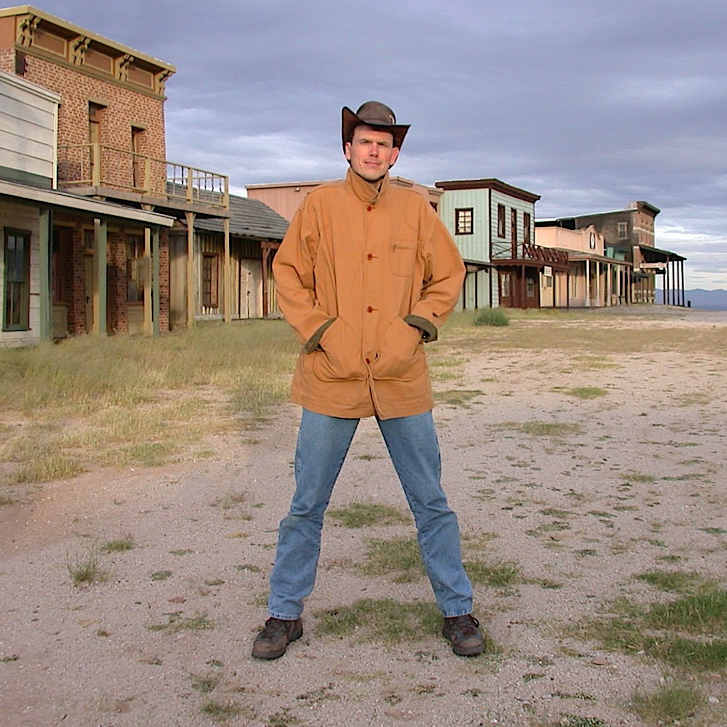 A man standing with legs apart, wearing a cowboy hat, tan jacket, and jeans in front of old western buildings in a desert landscape.