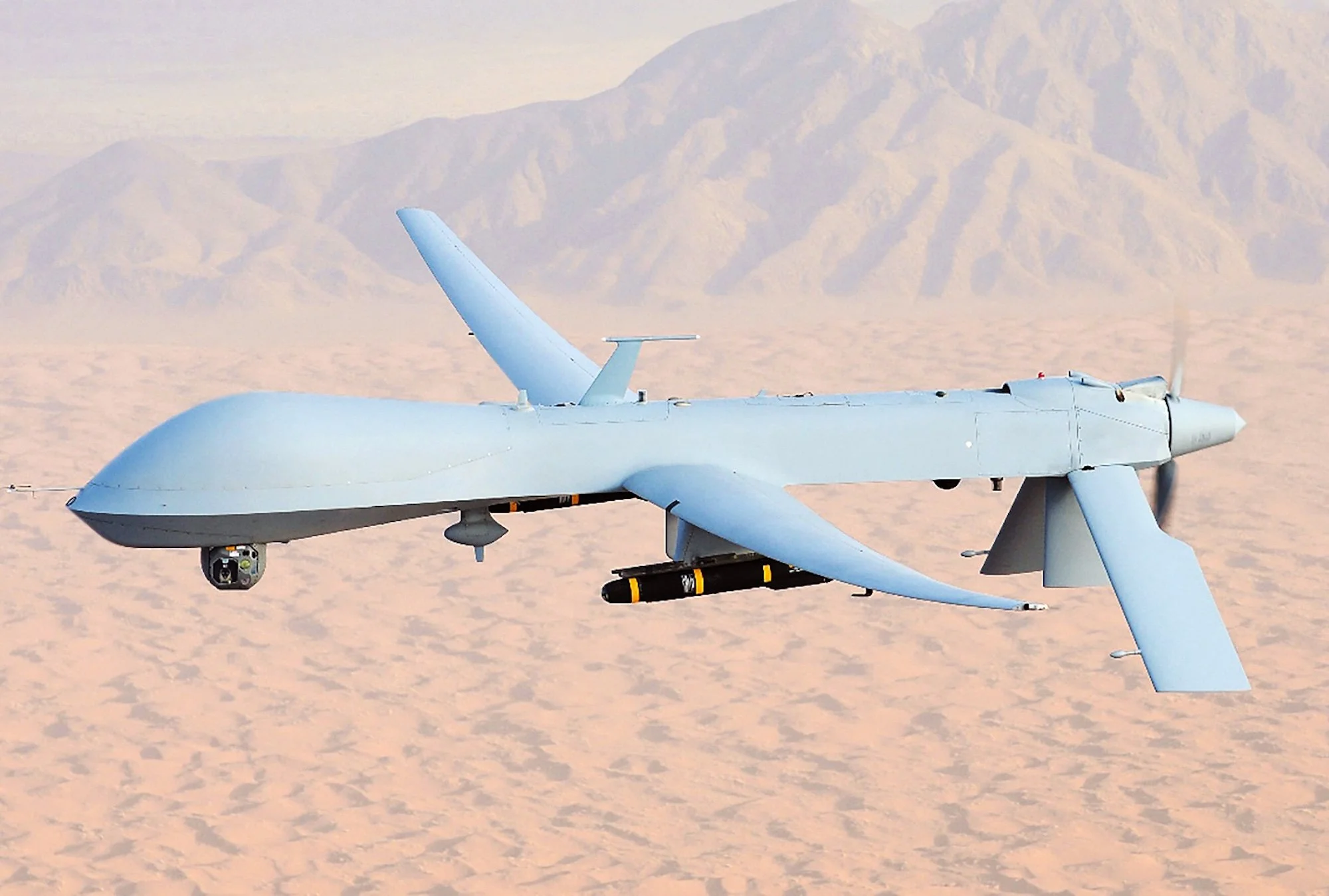An unmanned drone flying over desert landscape with mountains in the background.