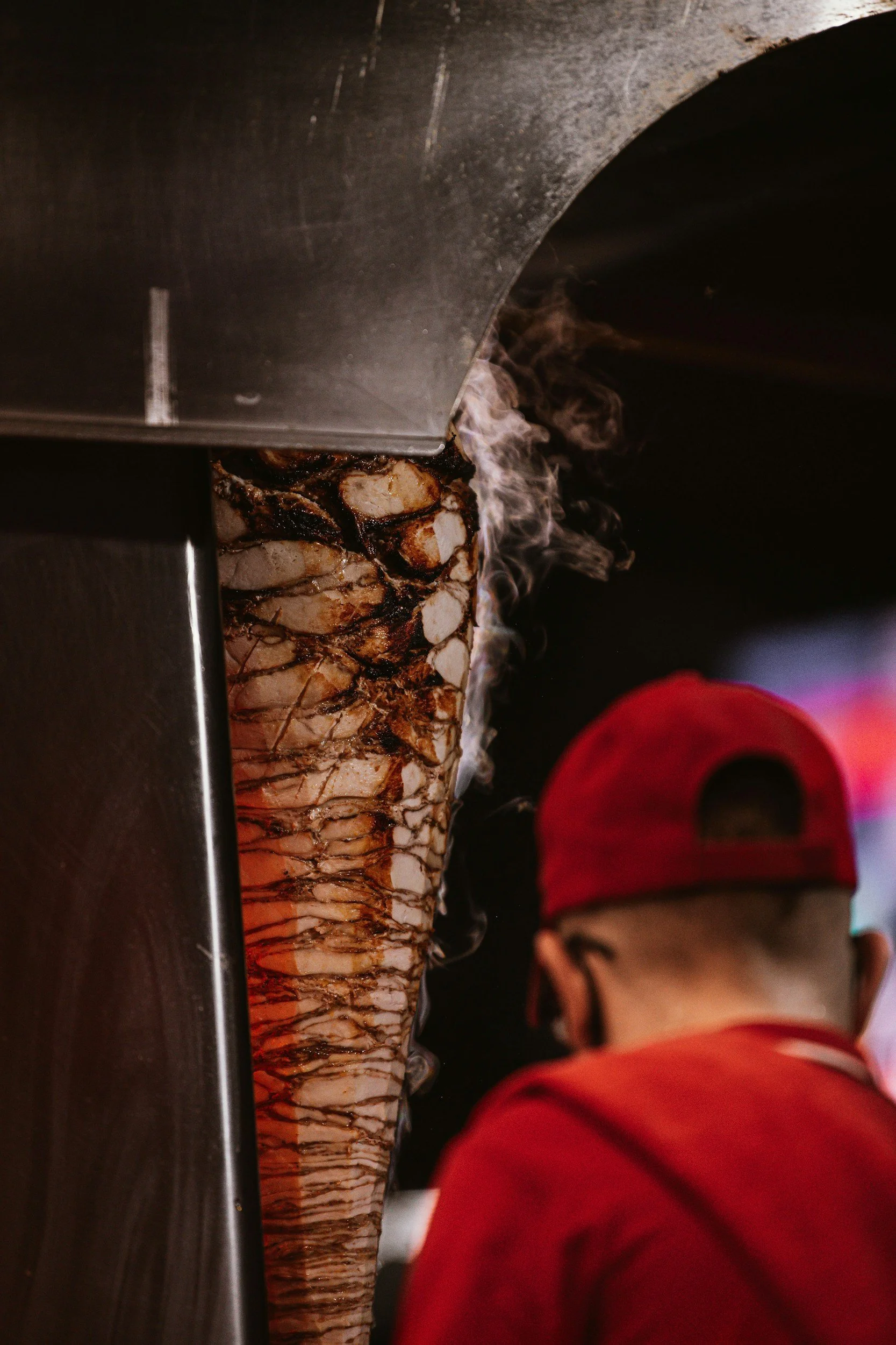 Close-up of a vertical rotisserie with slices of meat cooking, smoke rising from the heat, and a person in a red shirt in the background.