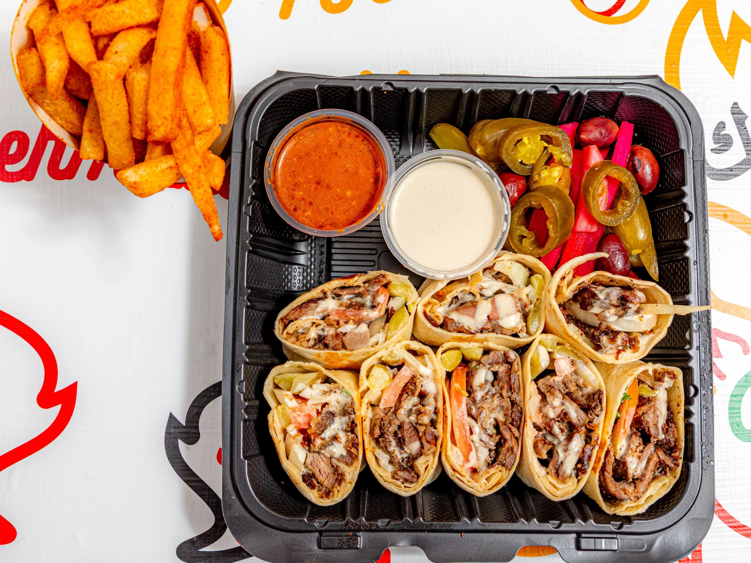 Tray of six beef and bean burritos with melted cheese and toppings, two small cups of creamy and red sauces, and a bowl of seasoned potato wedges with a side of jalapenos and pickled vegetables.