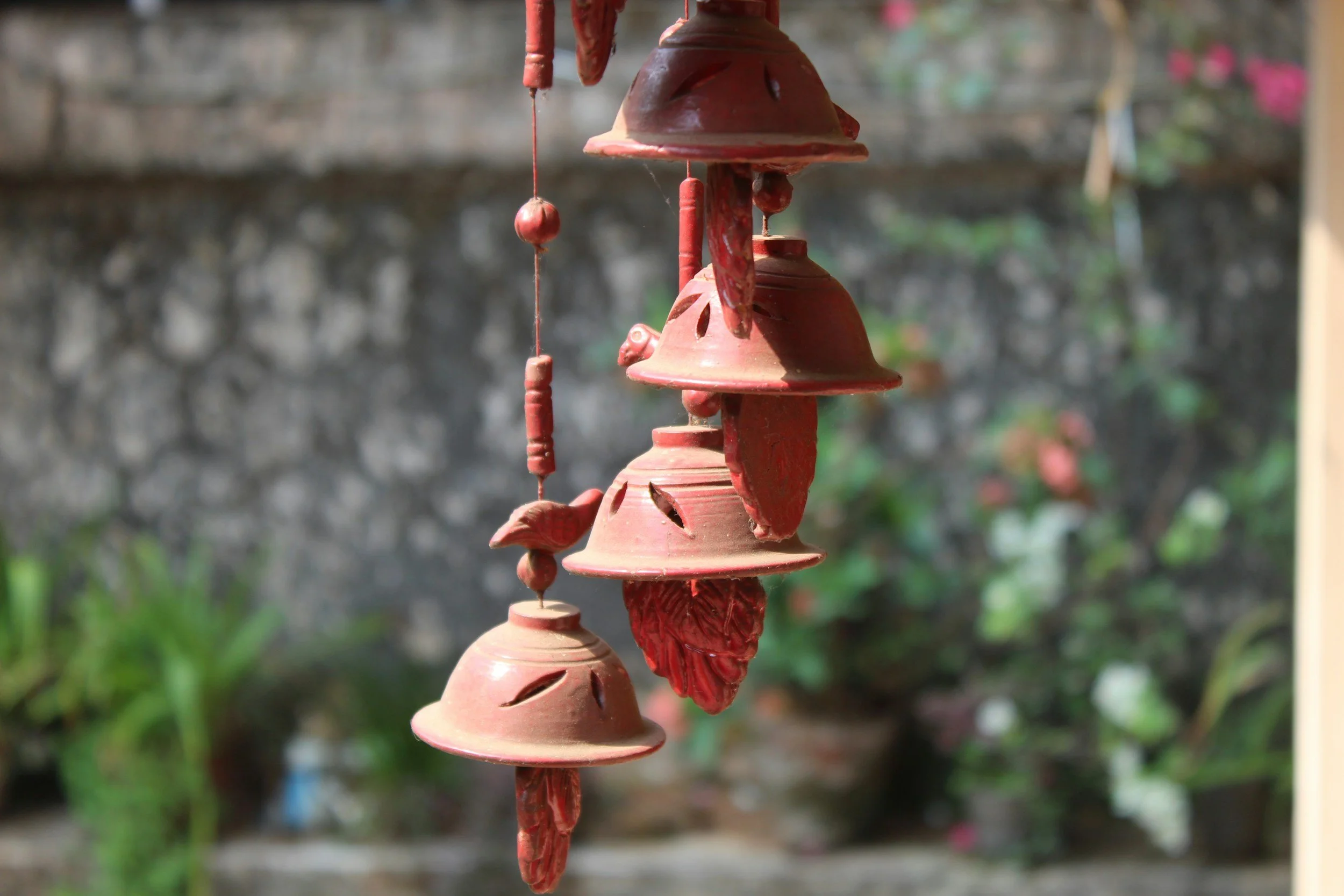 A hanging mobile made of small, reddish, clay-like wind chimes with leaf and bell shapes, hanging outdoors against a blurred natural background.