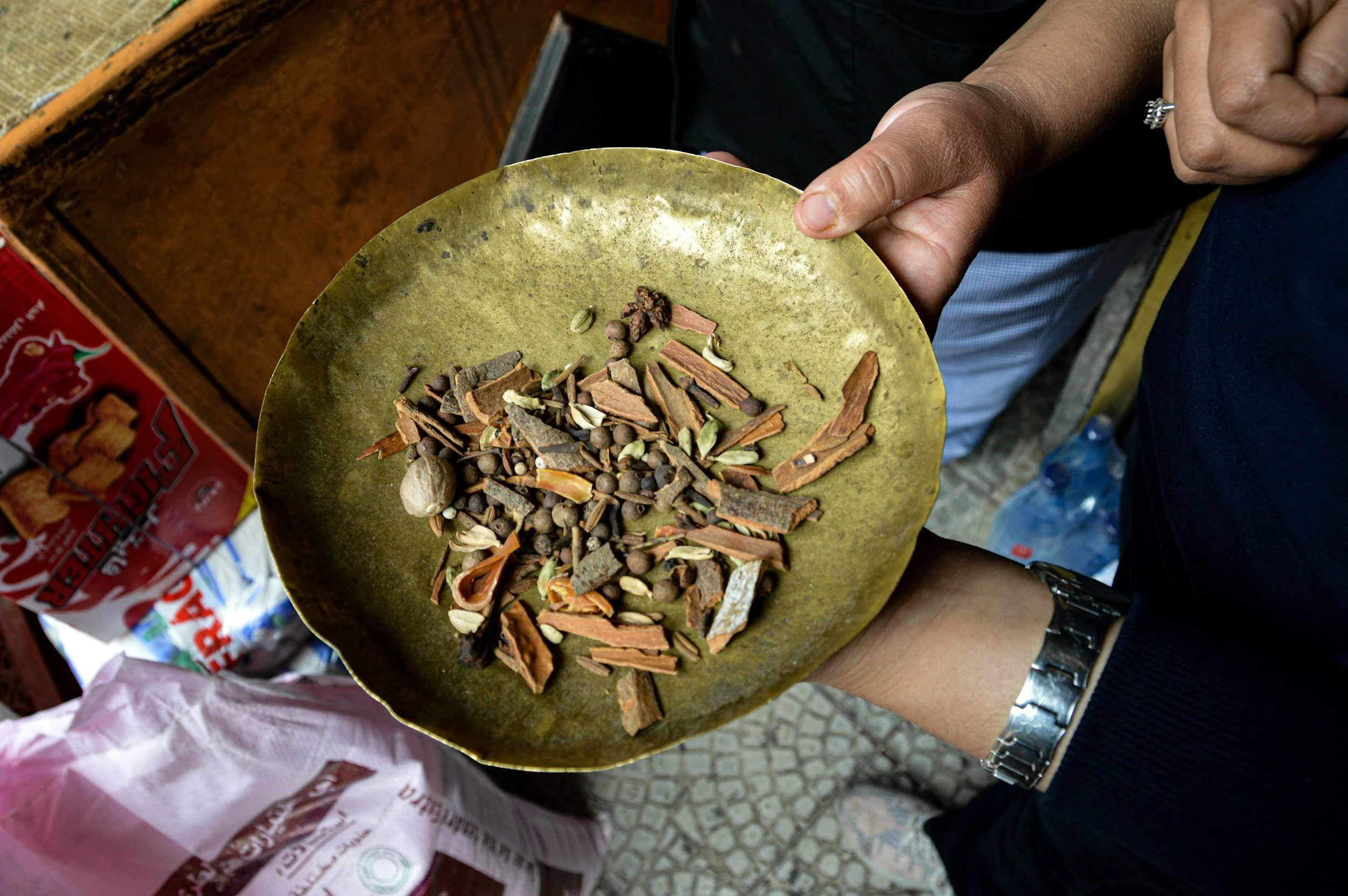 Person holding a yellow plate filled with various spices and dried herbs. Part of a red and white bag is visible in the background, likely containing more spices or herbs.