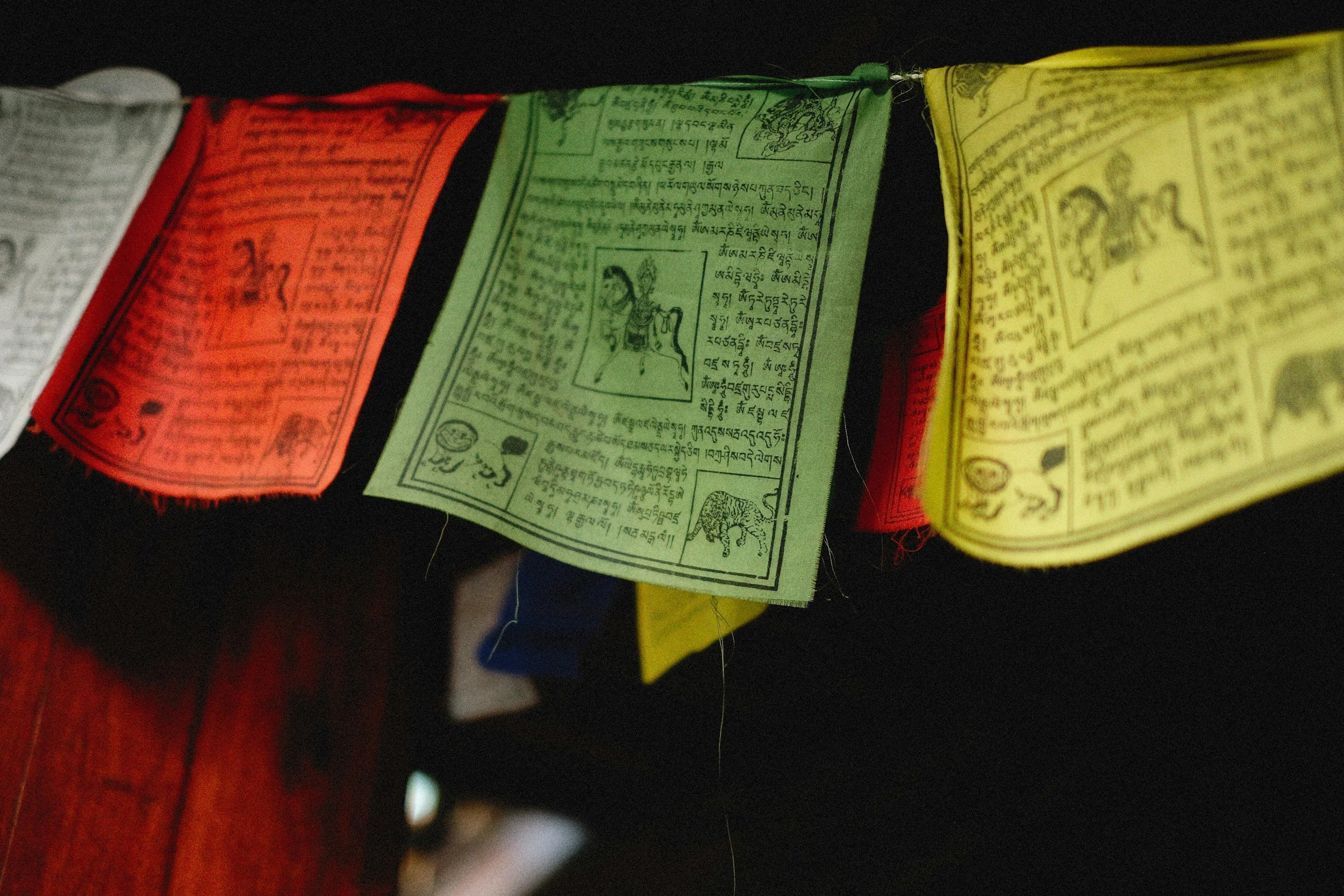 Close-up of colorful prayer flags hanging, with text and images in a script, possibly Tibetan or Sanskrit, on each flag.