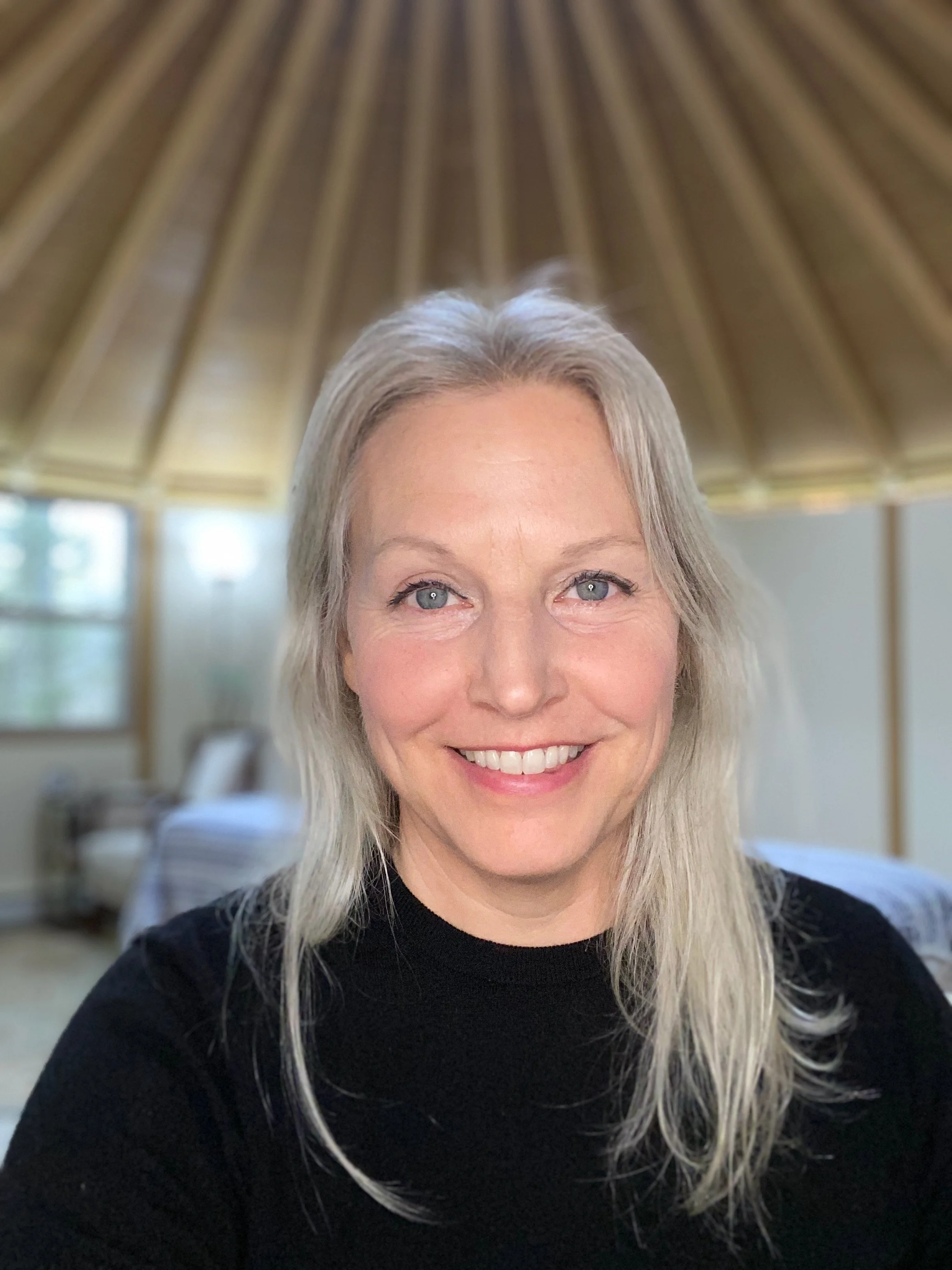 A woman with long, gray hair and blue eyes smiling at the camera in a room with a high, wooden vaulted ceiling.