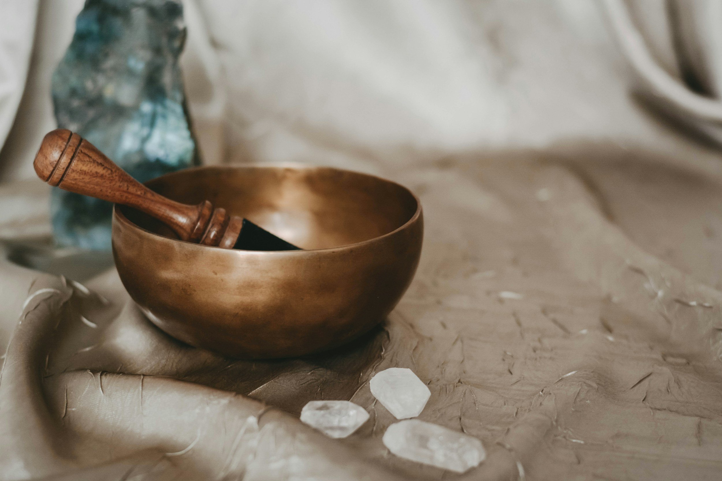 A small wooden singing bowl with a wooden striker inside, placed on a textured beige surface with scattered white stones, and a blurry glass bottle in the background.