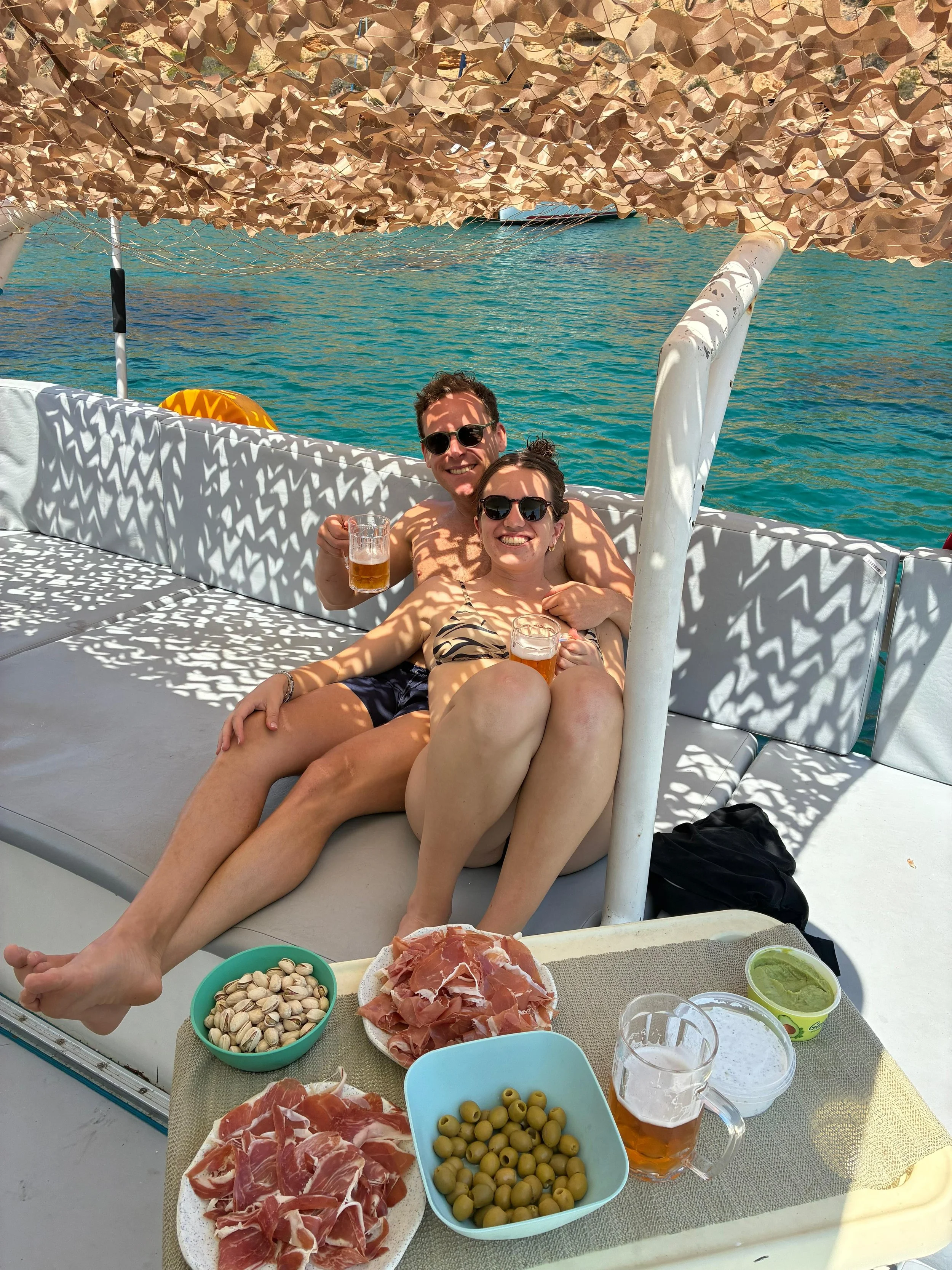 Two people are relaxing on a boat during daytime, enjoying drinks and food by the water. The table in front has bowls of olives, prosciutto, and green dip, with glasses of beer. They are smiling and wearing sunglasses, with a scenic body of water in 