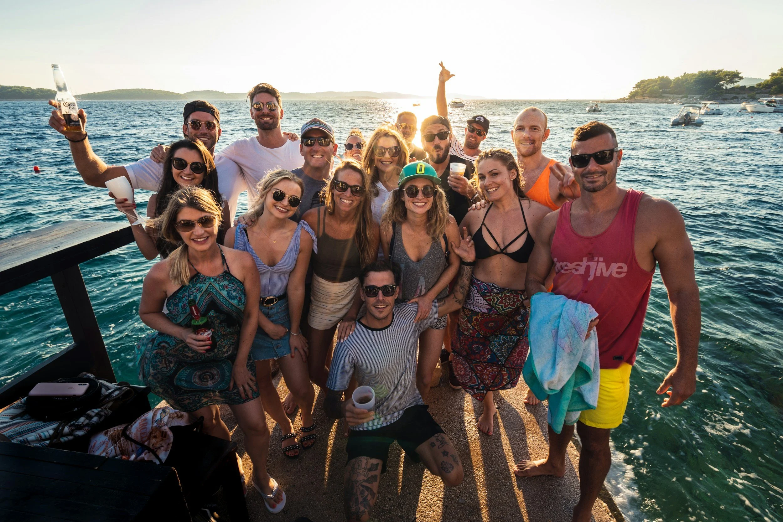 A group of friends on a boat dock by the water, smiling and enjoying drinks during sunset, with boats and distant land in the background.