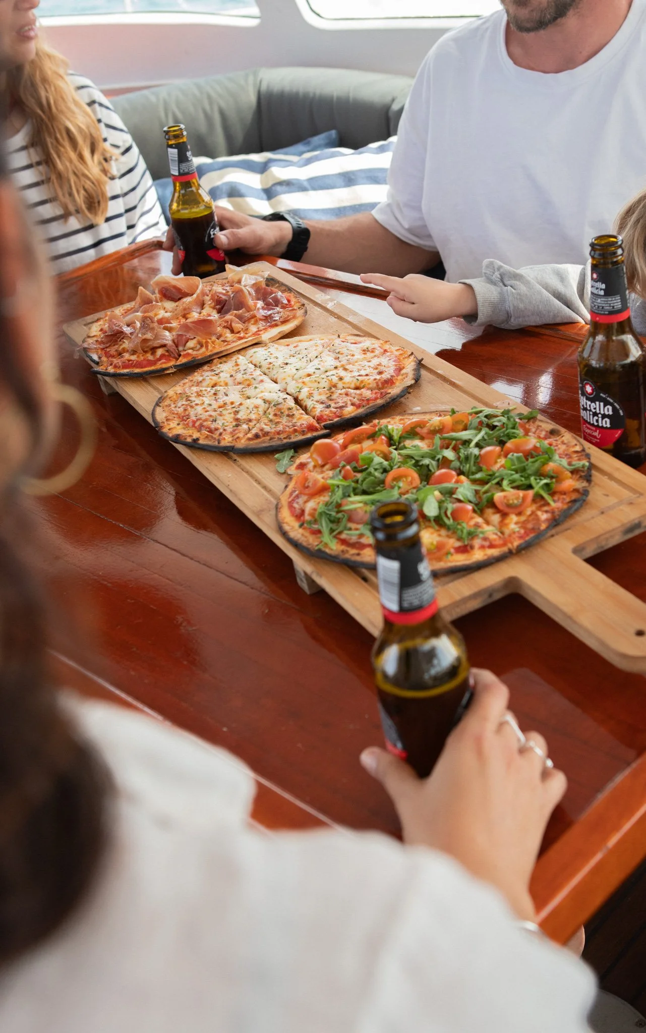 Close-up of children's hands reaching for pizza and holding beer bottles on a wooden table with three pizzas and a green salad.