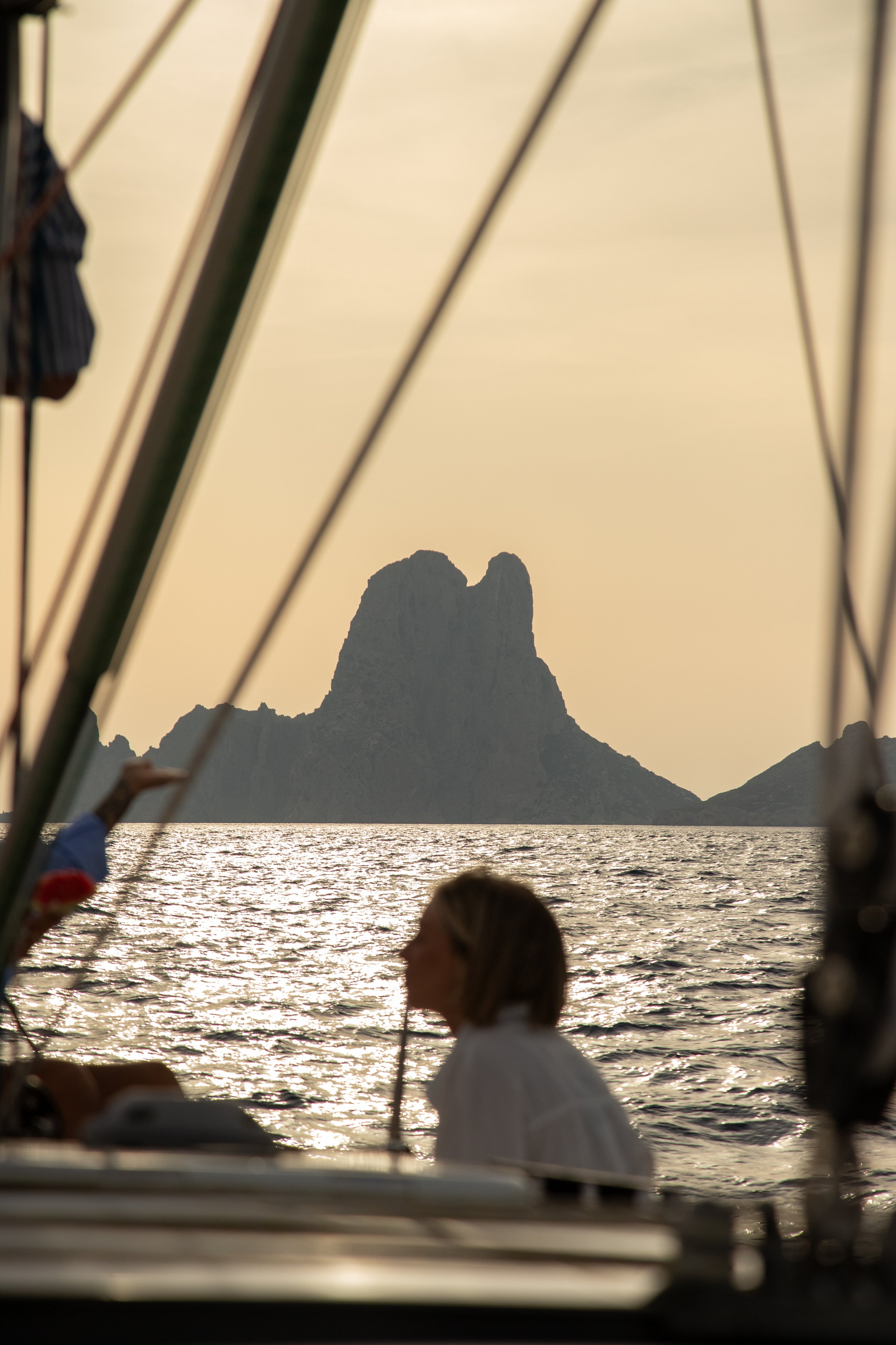 A photo taken from a boat showing a silhouette of a mountain or rock formation in the distance during sunset, with water in the foreground and parts of the boat and people on board visible.