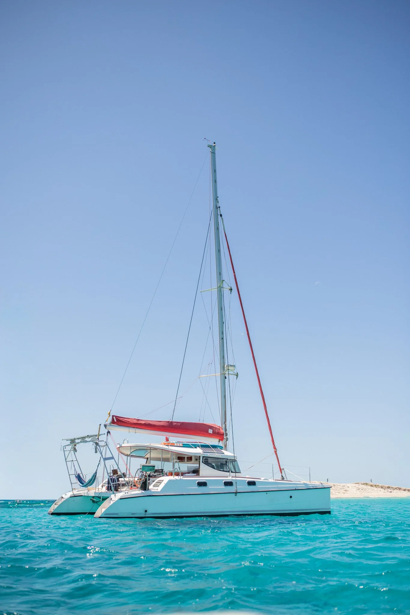 A white sailboat with a tall mast on clear blue water, a sandy island in the background under a blue sky.