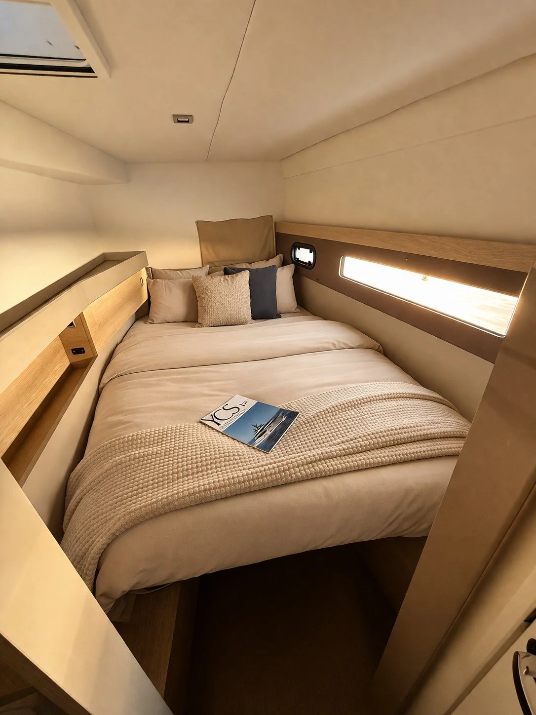 A cozy bedroom with a neatly made bed, decorated with pillows, a blanket, and a magazine on top. The room has wood accents, a small window, and a built-in shelf.