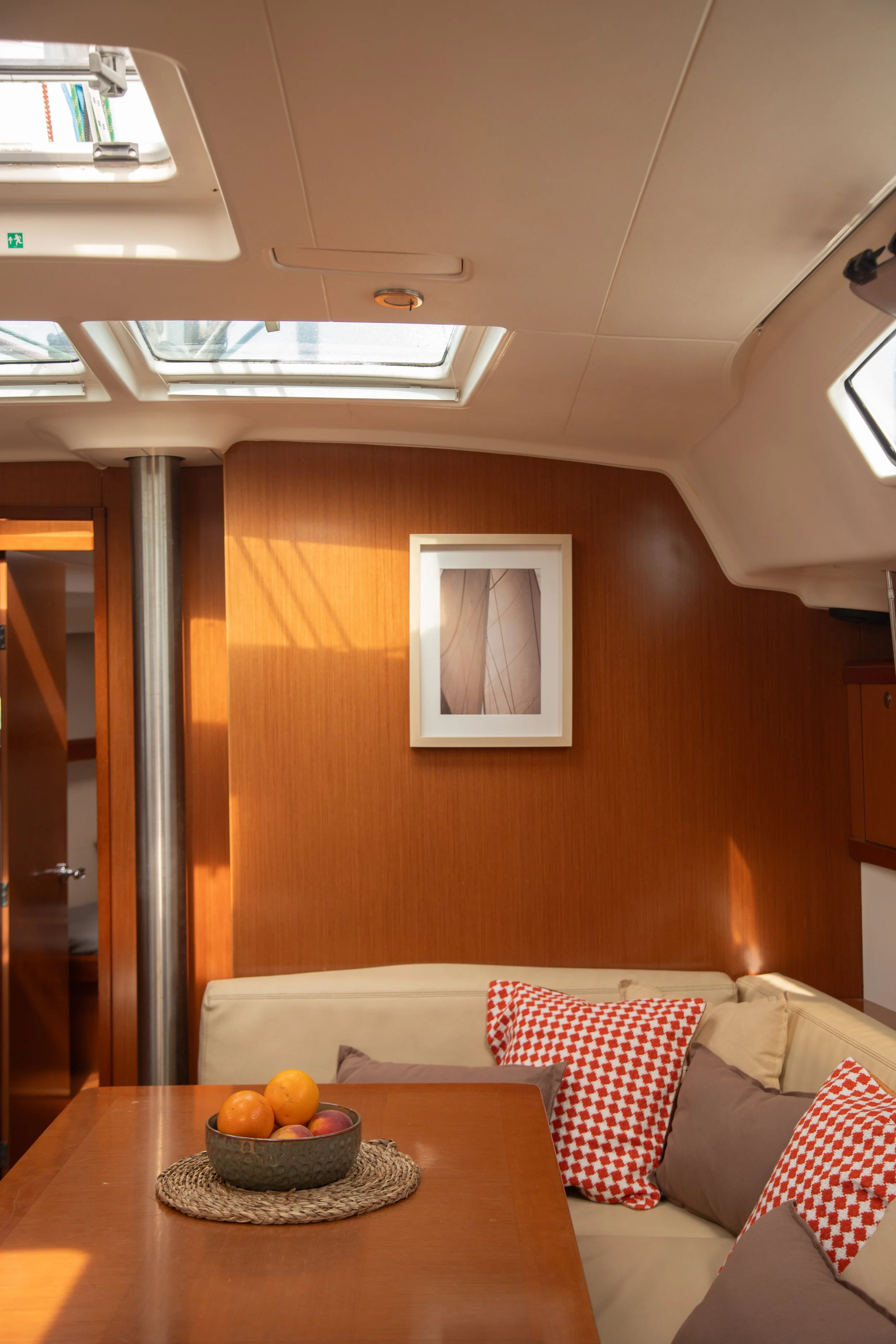A cozy indoor space with wooden walls, a dining table with a fruit bowl, and a beige sofa with red and white checkered pillows. The ceiling has skylights and a framed picture on the wall.