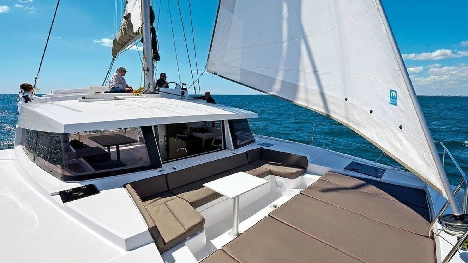 Luxury sailboat with tan cushioned seating area and white table on deck, under large white sails, on blue ocean under partly cloudy sky, with four people on the boat.