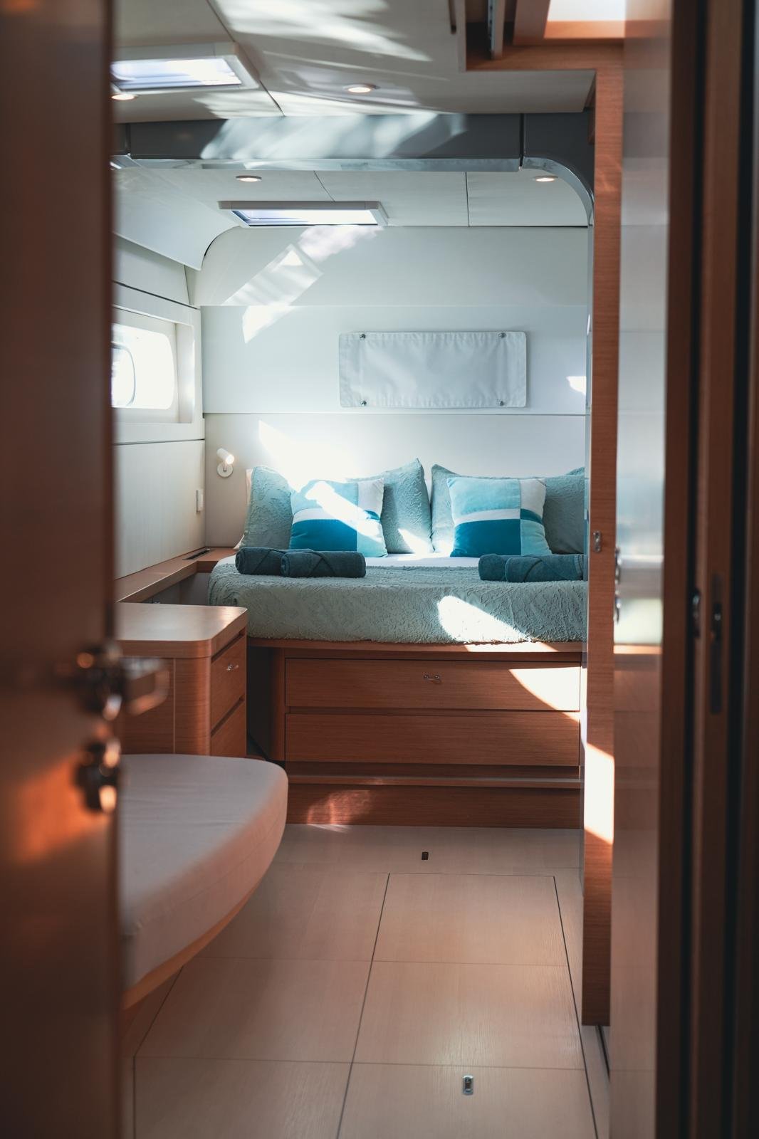 Inside the bedroom of a yacht with a bed featuring two rolled towels and blue pillows, sunlight streaming through a window, and a small overhead hatch.