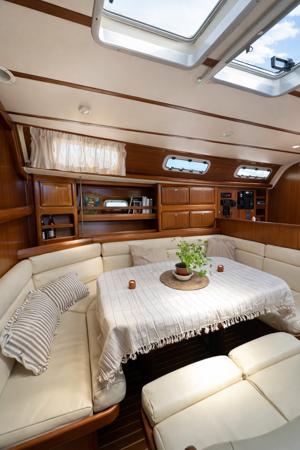 Interior of a boat or yacht with a cozy sitting area featuring cream leather cushioned seating arranged in a U-shape around a rectangular table with a striped tablecloth. A potted plant and small decorative candles are on the table. The background shows wooden cabinetry, a window with curtains, and portholes on the ceiling, with a skylight showing a partly cloudy sky outside.