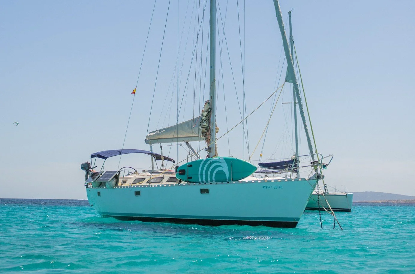 A white sailboat anchored in turquoise waters with a small surfboard on the side and a distant shoreline in the background.