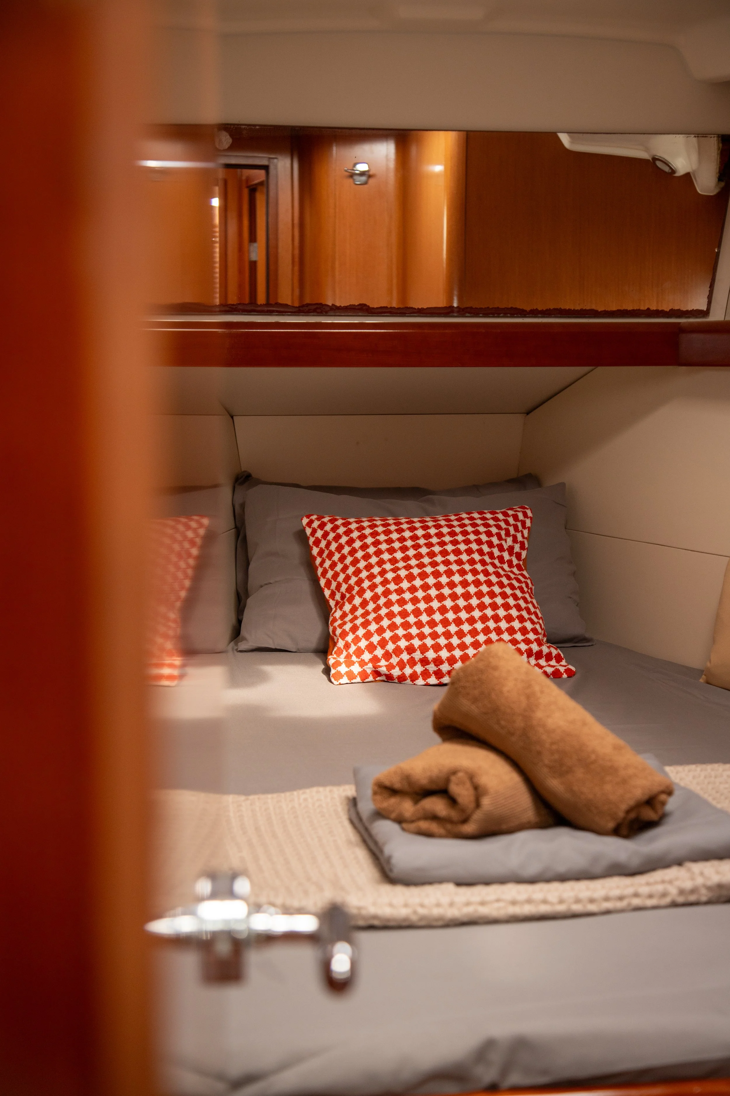 View of a neatly made bed with beige and red patterned pillows, a gray sheet, and neatly folded beige towels on top, inside a room with warm wooden walls and furniture.