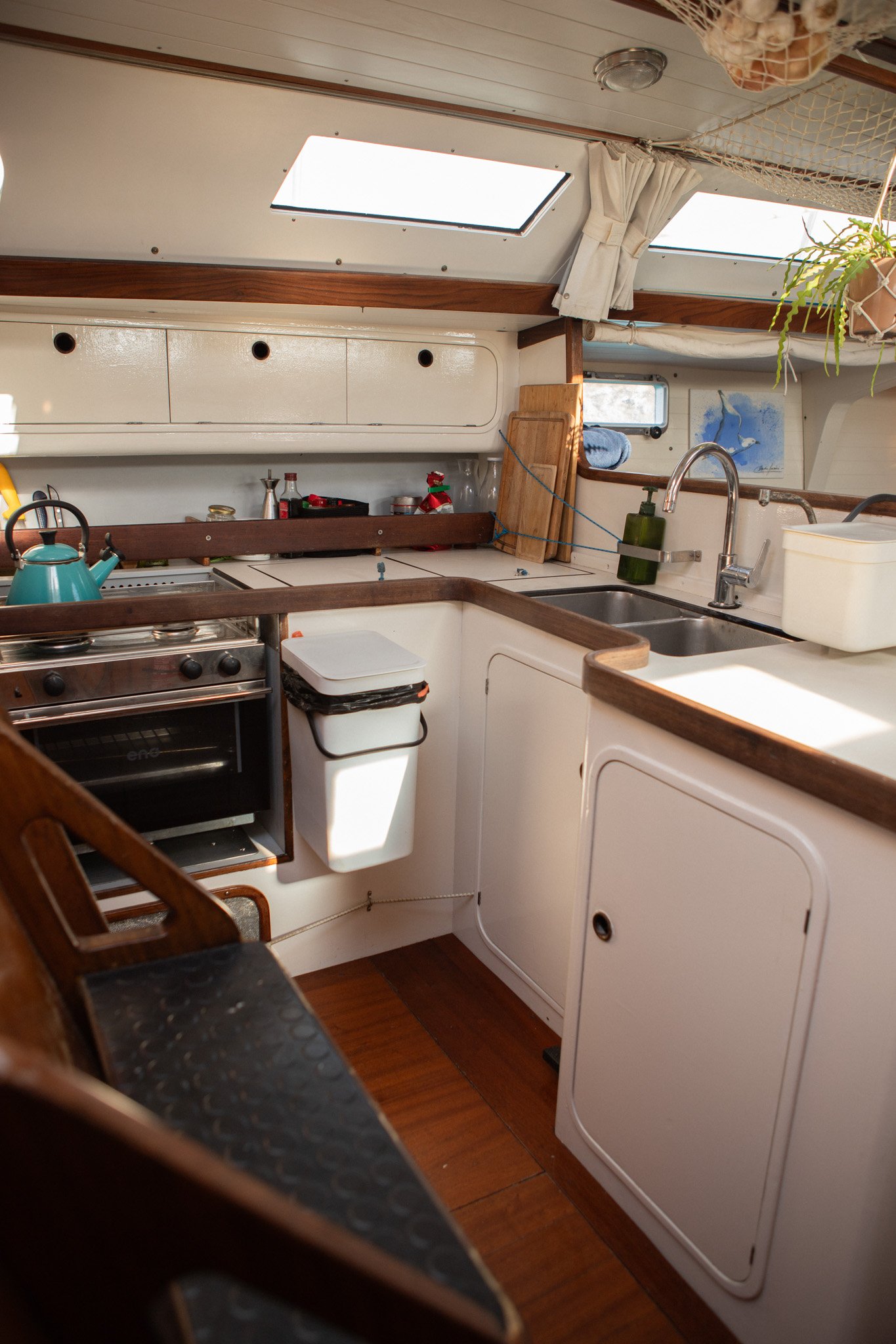 View of a compact boat kitchen with white cabinets, a small stove with an teal kettle, stainless steel sink, green soap dispenser, and various household items. Skylights and a window with white curtains are also visible.