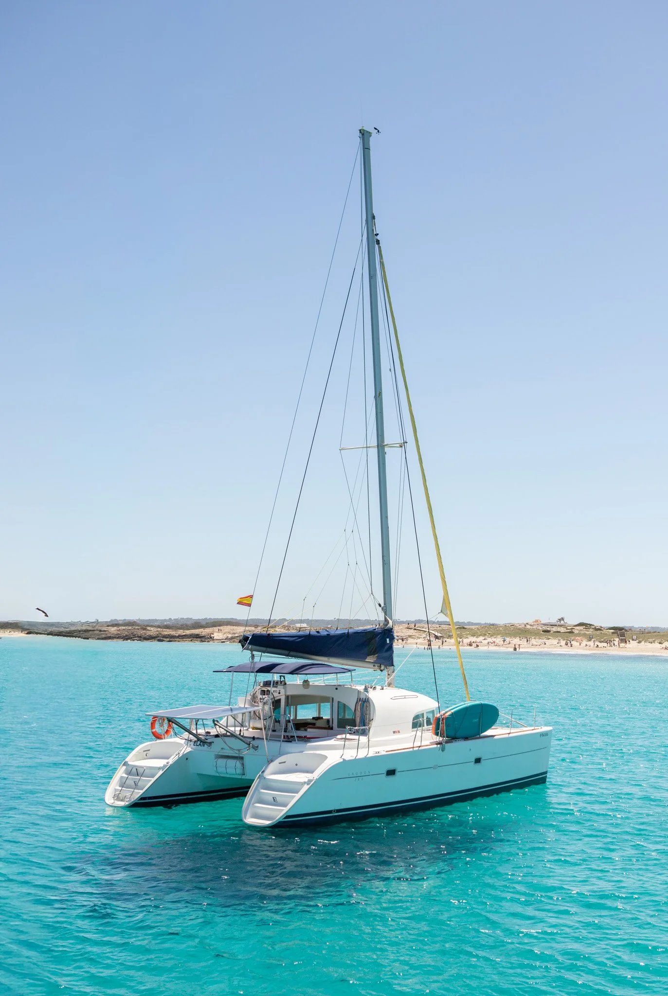 A white sailing yacht on turquoise water near a sandy beach with low hills and sparse vegetation in the background.
