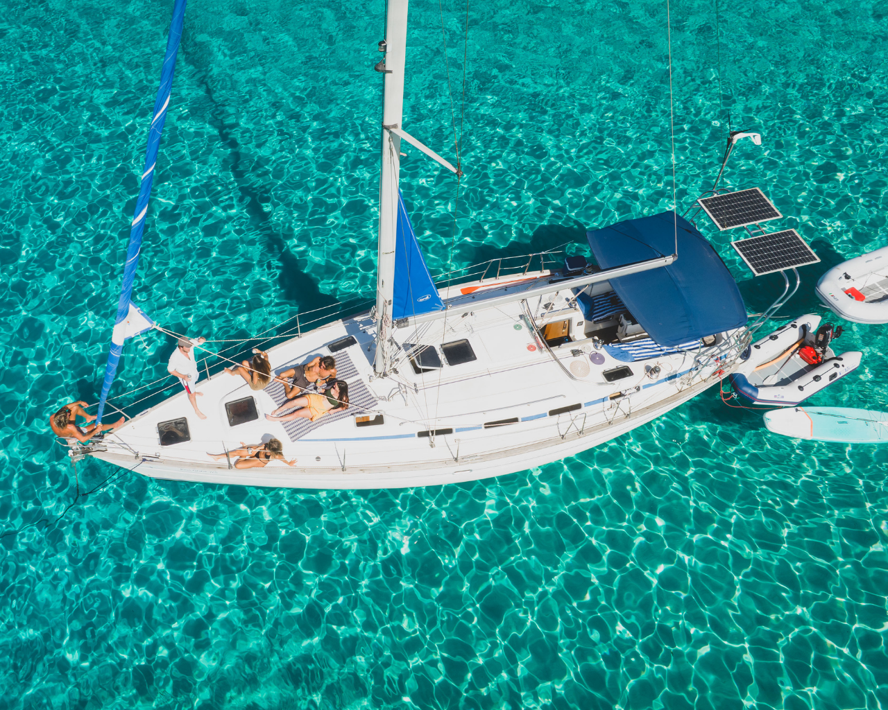Overhead view of a white sailboat with six people relaxing on deck, surrounded by clear turquoise water, with two small motorboats and a paddleboard nearby.