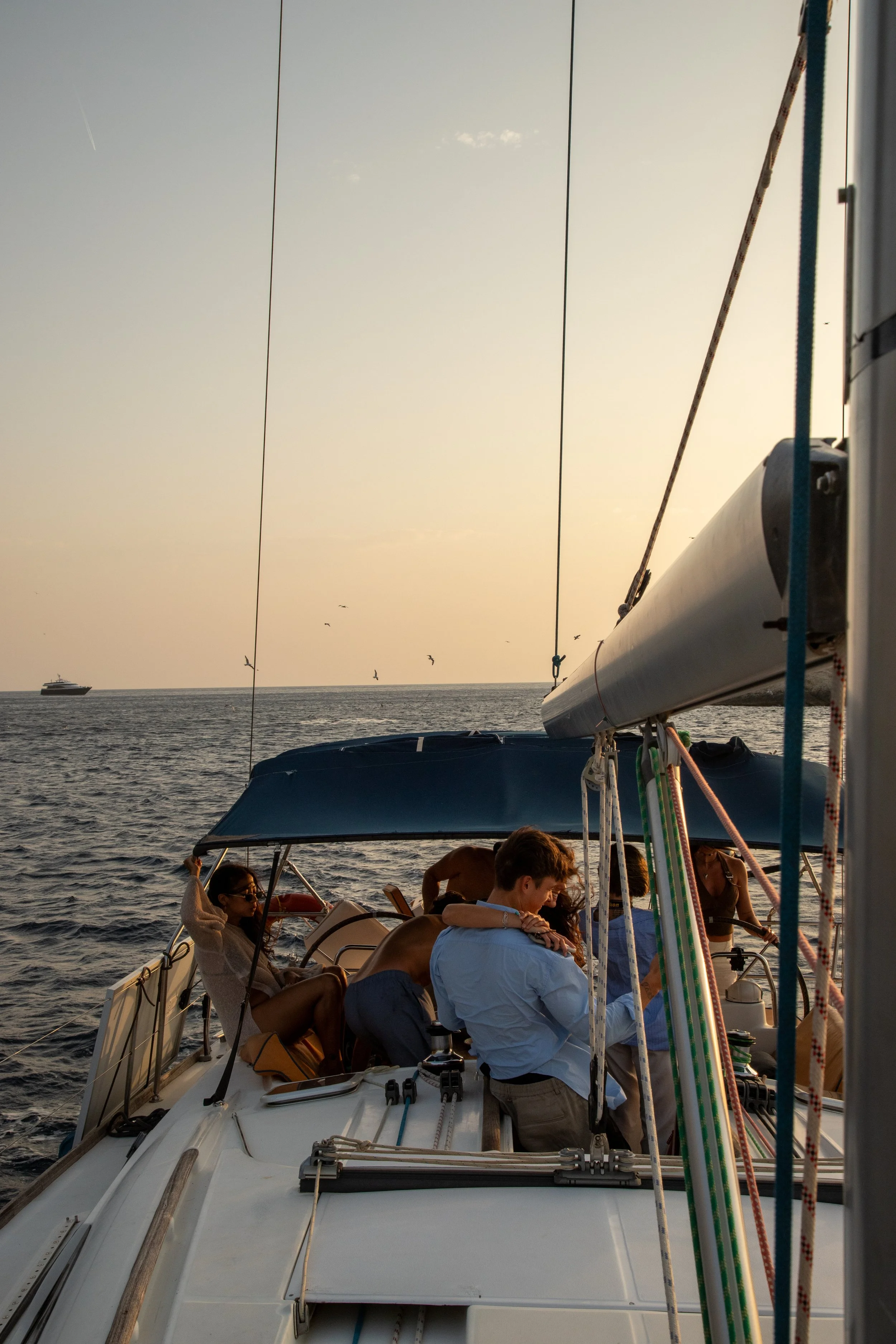 People sitting on a boat, some wearing sunglasses, with the sea in the background, and a boat sailing far away, during sunset