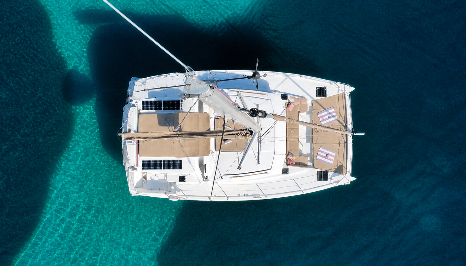 Overhead view of a white yacht with beige cushions on the deck, sailing on clear blue water.